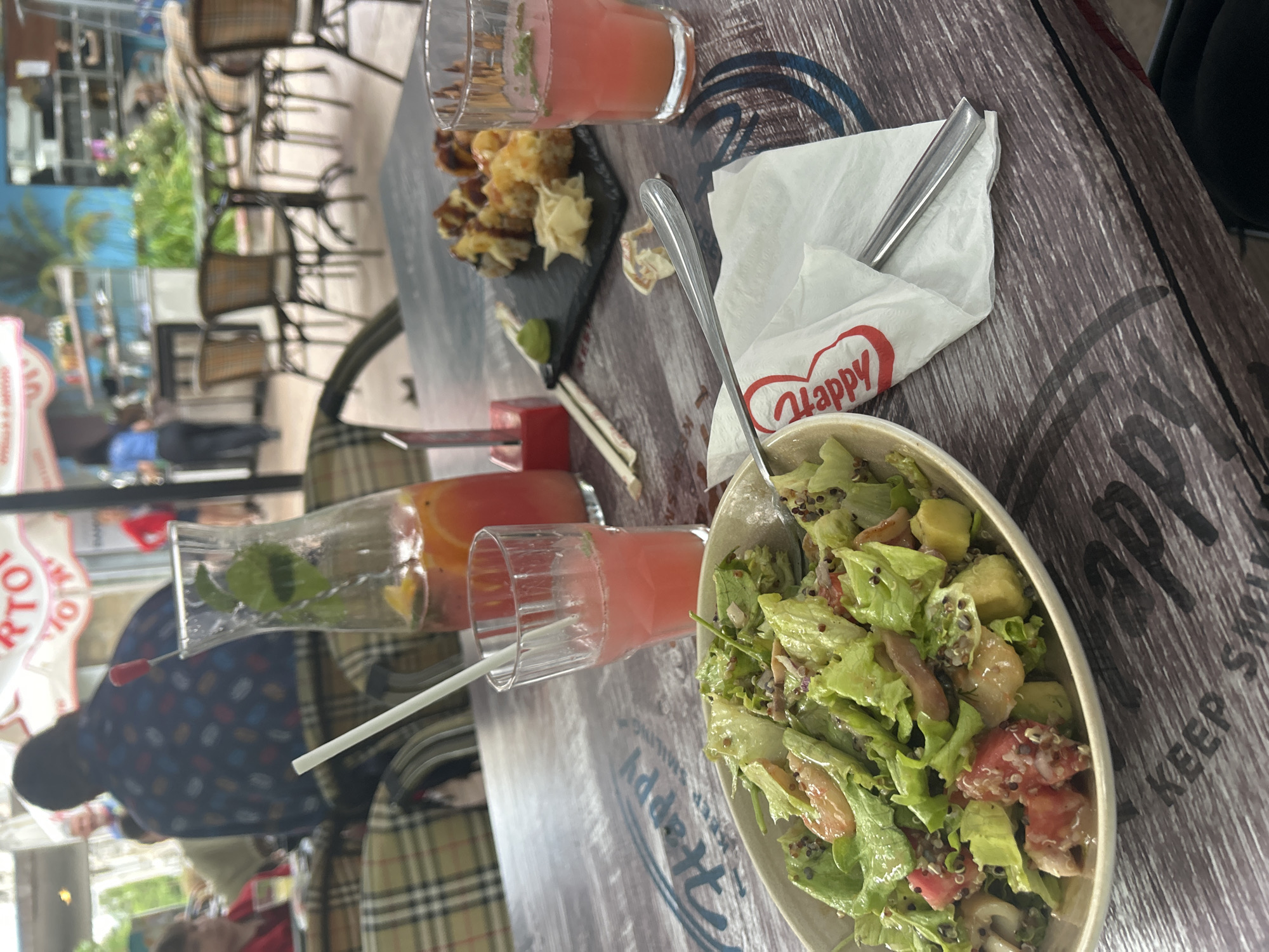 A photograph taken from a high angle shows a table at a restaurant, likely in Burgas, Bulgaria. The table is gray with a wood-like pattern. Present on the table are:
* A bowl of salad, with a spoon inside. The salad appears to contain lettuce, avocado, and tomatoes.
* A white napkin with the word "Happy" printed in red.
* A glass of a pink-colored beverage, possibly a cocktail.
* A small plate with sushi or similar bite-sized food items, along with chopsticks and a small mound of wasabi.
* Another glass of a pink-colored beverage with a straw.
* A taller glass with what appears to be a drink containing orange and lime.
Outside the restaurant, chairs and tables are visible, along with people walking by. The restaurant is likely located in an area with shops and greenery. A blurred person wearing a patterned shirt is visible in the foreground.