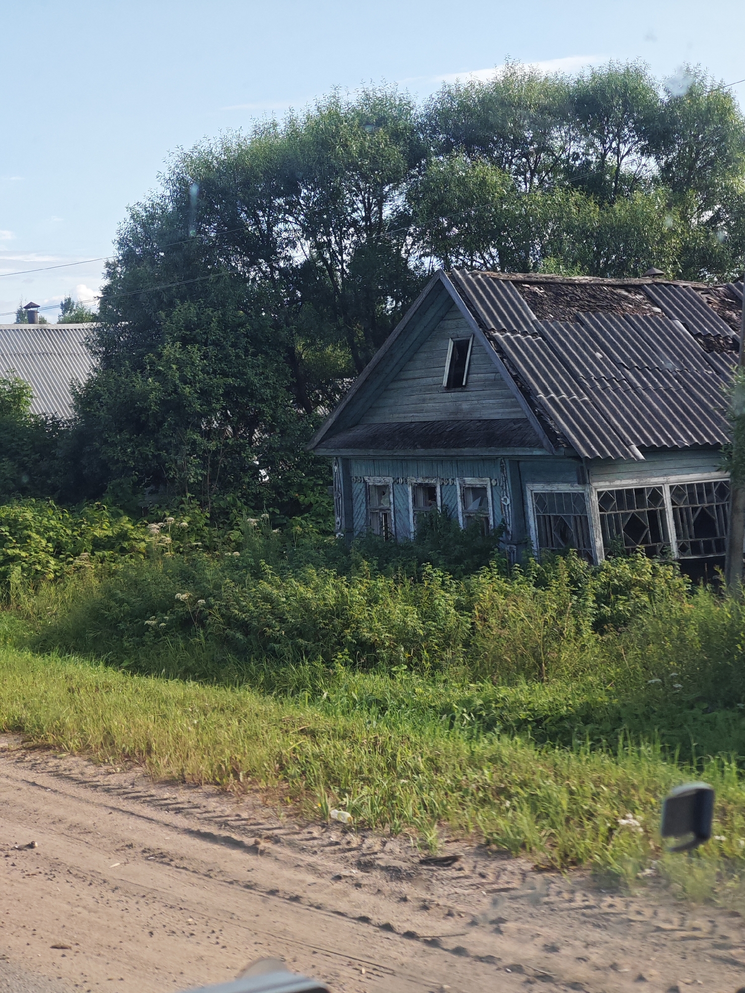 An abandoned, weathered, light blue wooden house stands amidst overgrown green foliage in the village of Budovo, Russia. The house has a dilapidated corrugated roof, a visible attic window, and multiple broken windows. A dirt road with tire tracks is in the foreground, with a grassy embankment separating it from the dense vegetation surrounding the house. Tall trees with lush green leaves form a dense canopy behind the structure, and a portion of another building with a grey, corrugated roof is visible to the left of the frame. The sky above is a clear light blue with a few faint white clouds.