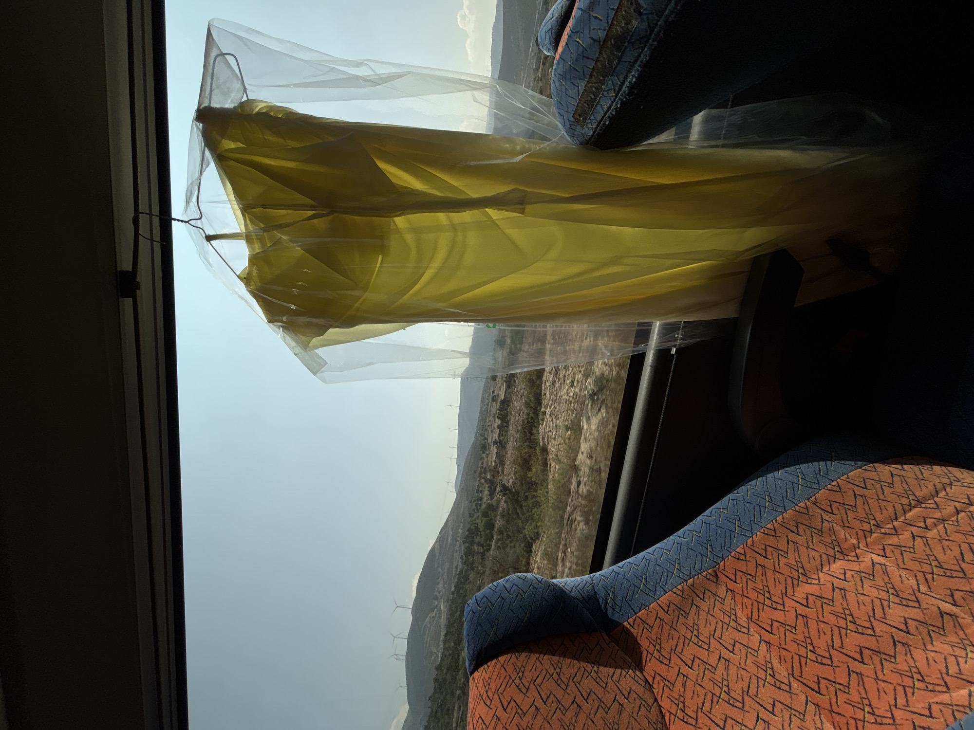 The scene is inside a bus traveling through Azumbilla, Mexico. The view is through the window. There is a yellow garment covered in plastic hung on a clothing hanger. Outside the window there is a landscape of hills with green foliage. There are also visible wind turbines. Two patterned bus seats are also visible.