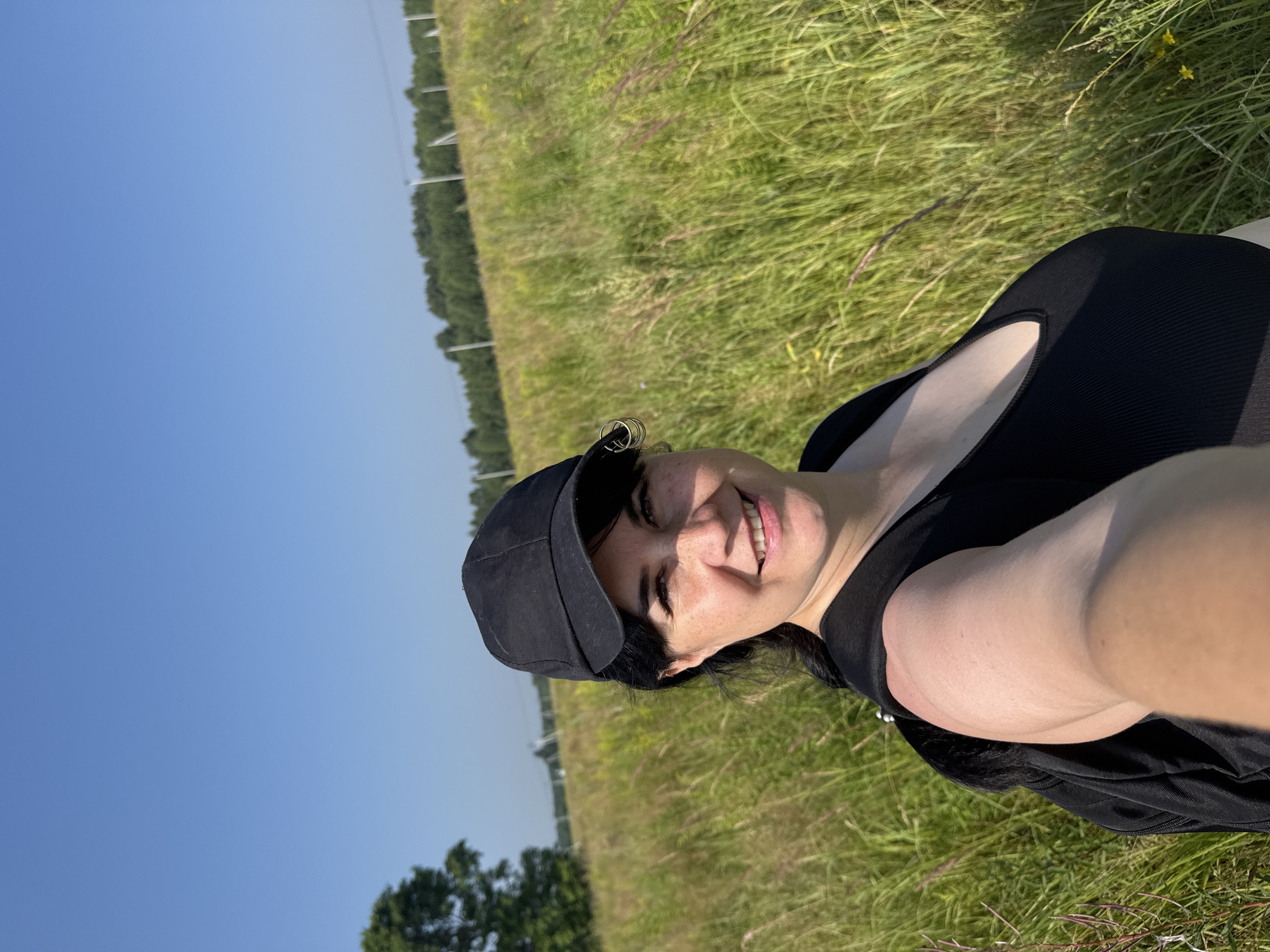 The image shows a woman in a field on a sunny day. She is wearing a black baseball cap and a black tank top, and has a backpack on. She is smiling and looking at the camera. The field is filled with tall green grass and some yellow flowers. In the background, there is a line of trees and a clear blue sky. Based on the context provided by the prompt, this scene is located in the city of Izhevsk, Russia. There are no other individuals present.