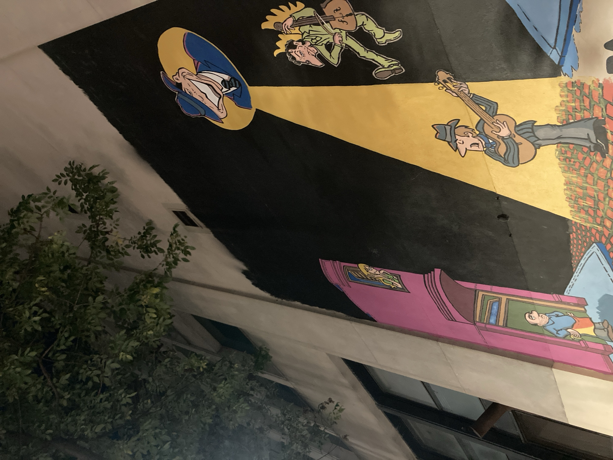 The image displays a colorful, cartoon-style mural painted on a building wall, captured at an oblique angle that makes the artwork appear rotated counter-clockwise. The scene depicts a night or dusk setting, with a large, dark green leafy tree partially obscuring the lower left foreground.
The mural features several characters under prominent yellow spotlights against a dark background. On the left, a man in a fedora and blue trench coat looks upwards into a circular spotlight. To his right, a man with a distinctive long nose, dressed in a light green suit, energetically plays a violin, illuminated by a strong yellow beam. Further right, another character wearing a blue hat and striped shirt plays an acoustic guitar, also bathed in a yellow spotlight. Behind these figures, a backdrop resembling a red brick wall and a large blue, wavy shape are visible.
Below the musicians, the mural extends to a vibrant pink building facade with decorative elements. A balding man in a blue shirt and yellow apron is shown peering out from a window in this pink structure. Above this building, a small detail reveals another character lying on what appears to be a flying carpet. The art style is bold and cartoonish, utilizing strong outlines and bright, contrasting colors. No visible text is present anywhere in the image.