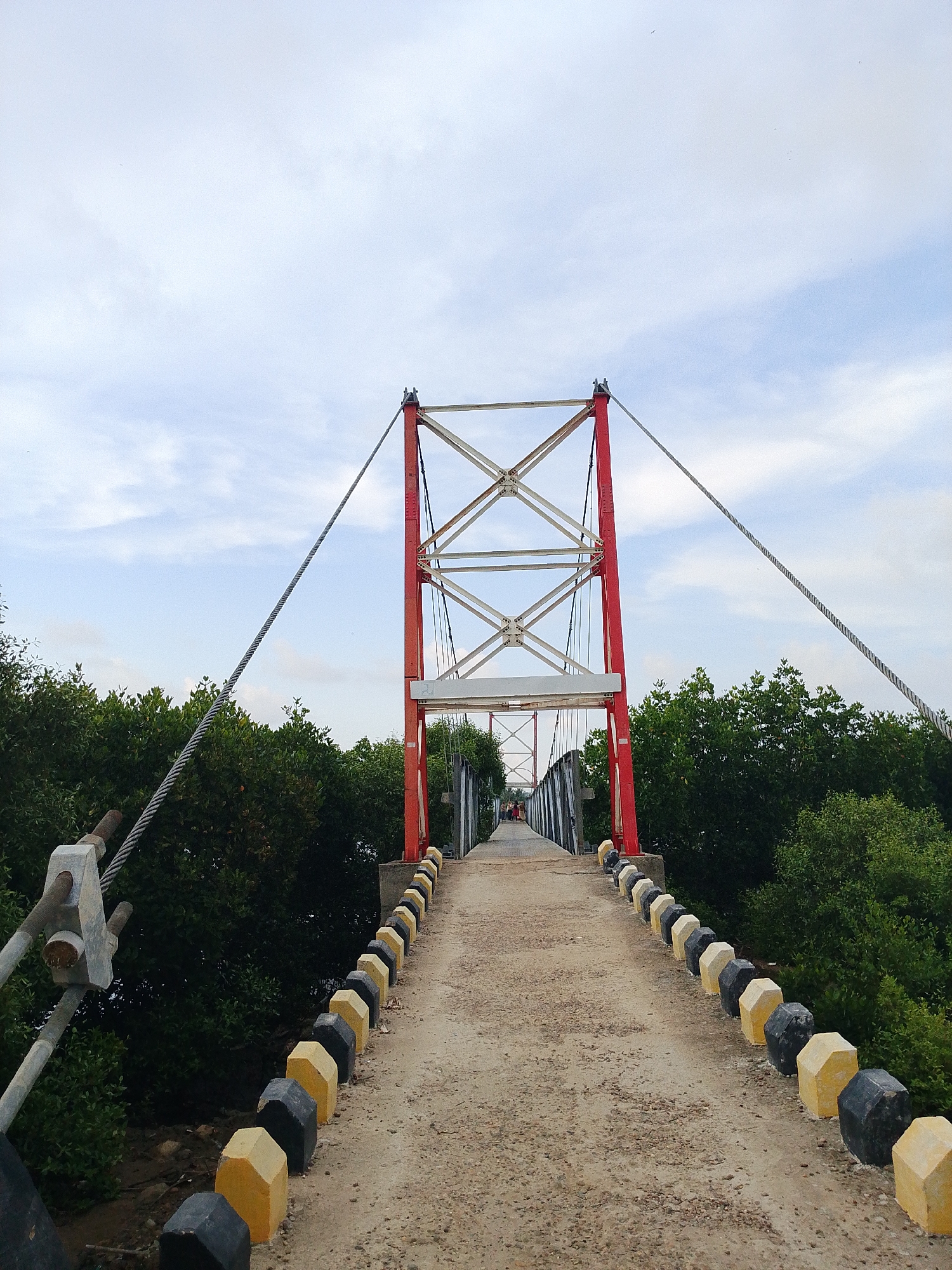 The image depicts a bridge in Lhokseumawe, Indonesia. The bridge is a suspension structure with red and white towers. The walkway is lined with alternating yellow and black hexagonal concrete barriers. Steel cables support the bridge, extending from the towers to the bridge deck. Lush green trees flank both sides of the bridge, and the sky is overcast with light clouds. In the distance, figures appear to be walking on the bridge.