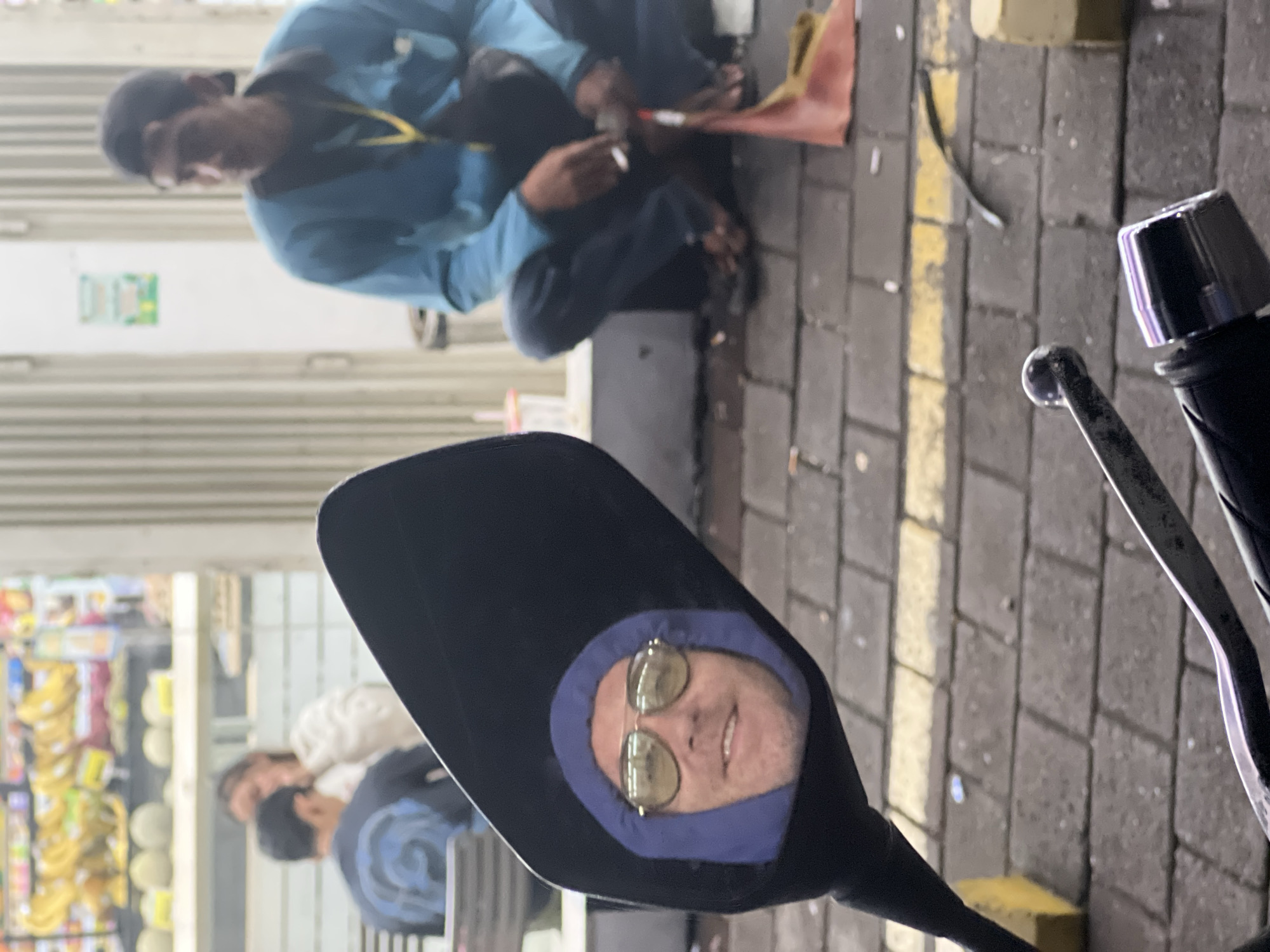 The image is taken from the perspective of a person on a motorcycle, looking into the motorcycle's side mirror. The mirror reflects the person taking the picture; they are wearing sunglasses and a blue hooded jacket.
In the background, slightly out of focus, there are other people: one person is crouched down near the curb, apparently working on something or picking something up, and another two people are sitting in front of a store. The store appears to be a small convenience store, with shelves full of various goods visible through the glass doors.
The street is paved with gray bricks, and there is a yellow painted line marking the edge of the road. Debris litters the street, including some garbage and a small piece of fabric.
The location appears to be in the city of Denpasar, Indonesia.
