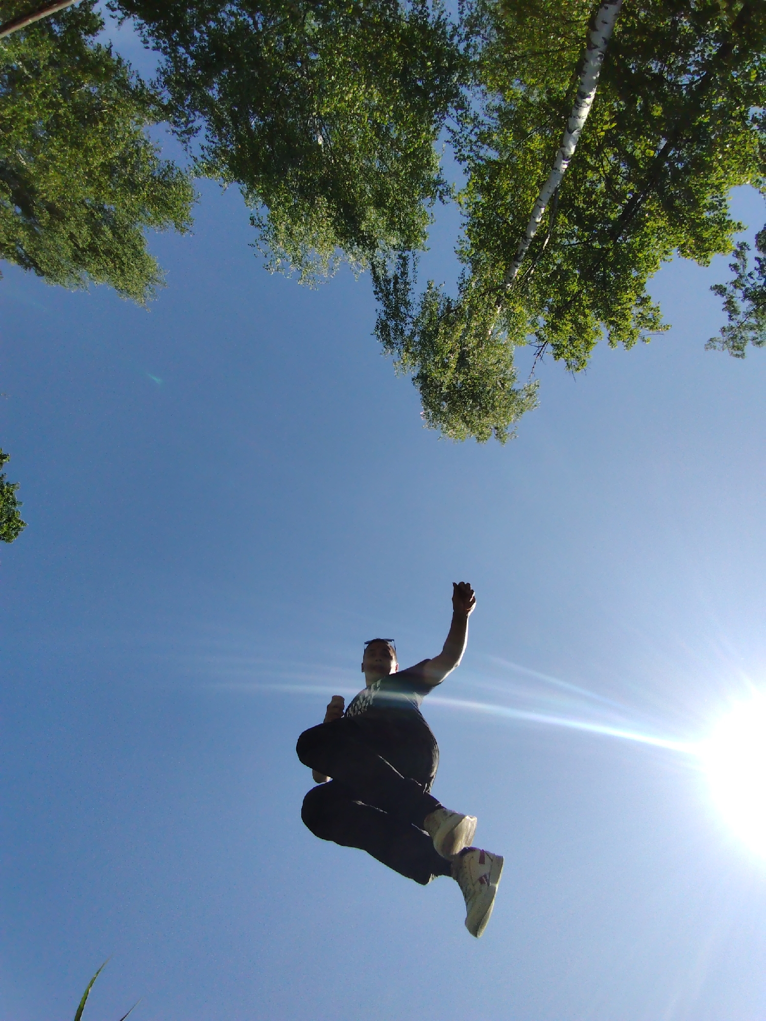 A low-angle shot captures a person in mid-air against a clear blue sky with trees visible in the upper part of the frame. The person appears to be leaping or jumping, with one arm raised. The sun is shining brightly, creating a lens flare effect. The location is Chaikovsky, Russia.