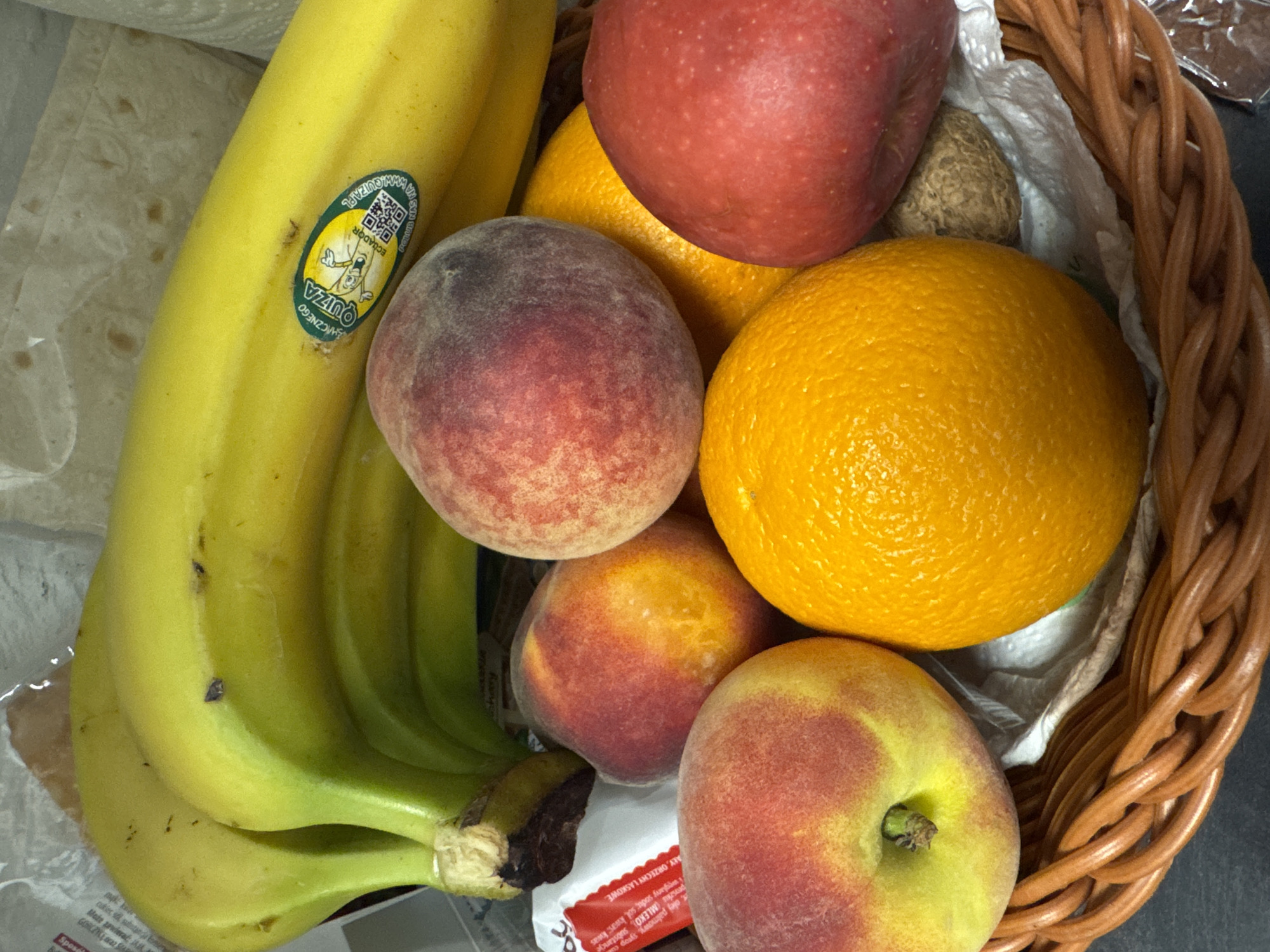 The image shows a basket overflowing with fresh fruit, including a bunch of bananas with a "QUIZA ECUADOR" sticker, two peaches, two oranges, and one red apple. A single walnut rests among the fruit. The basket itself is made of woven brown material and sits on a dark surface. In the background, there are glimpses of packaging materials, one of which has Polish text visible. The scene appears to be an indoor setting, possibly a kitchen or pantry, and there is no indication of any specific location in Poznan, Poland. There are no people or actions depicted in the image.