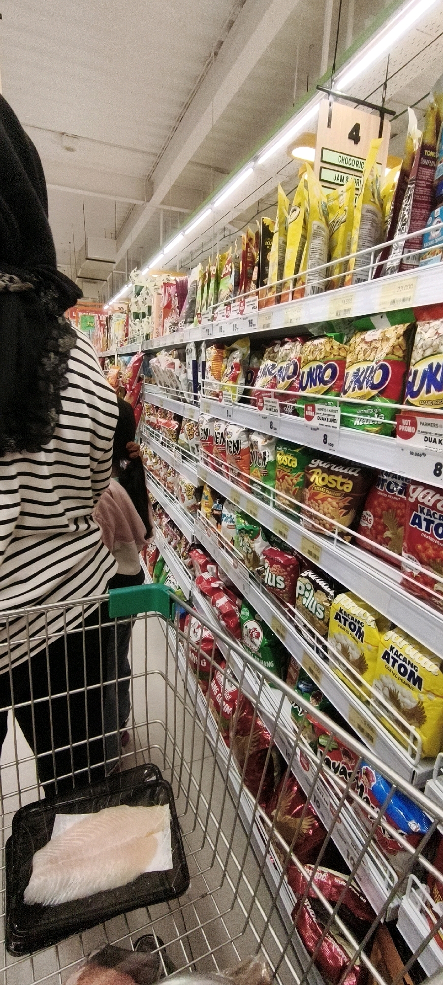 A person wearing a black hijab and a black and white striped shirt is pushing a metal shopping cart through a brightly lit supermarket aisle in West Jakarta, Indonesia. The cart contains a black plastic container with two pieces of raw white fish on a white paper lining, positioned in the lower front of the cart. The shelves on either side of the aisle are fully stocked with a variety of packaged snacks, chips, and other food items, predominantly in colorful bags and boxes. Prominent brands and product names visible include "UNRO," "ROSTA," "PLUS," and "KACANG ATOM." Signage with prices and product information is displayed above the shelves. The overall environment suggests a typical grocery shopping scene within an Indonesian supermarket.