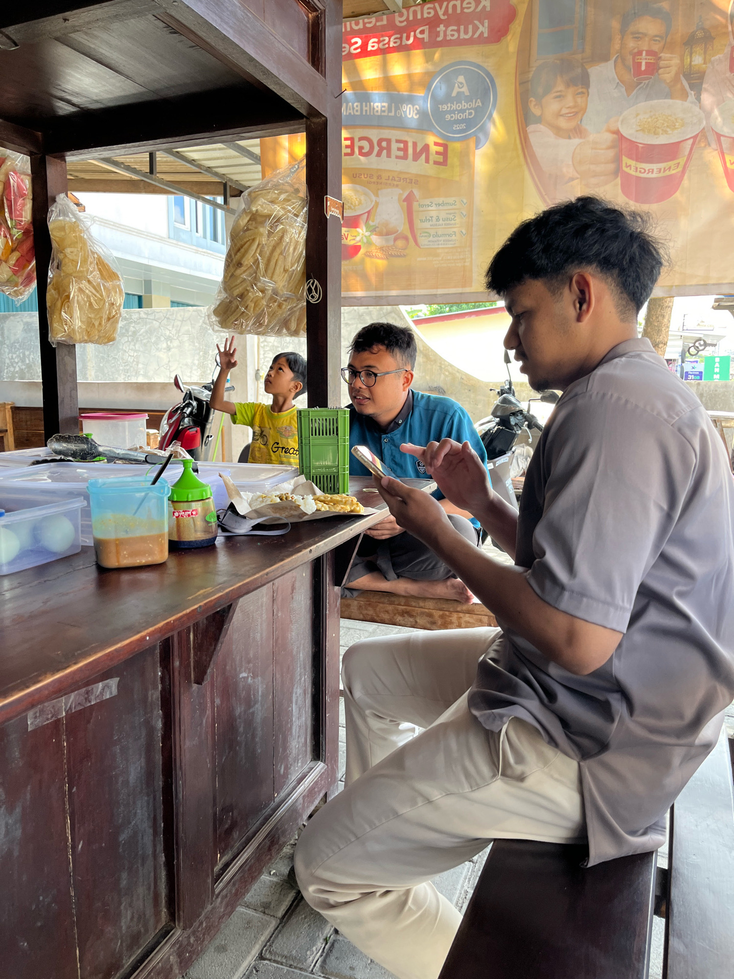 In Banguntapan, Indonesia, a roadside food stall displays bags of snacks, possibly crackers or chips, hanging in plastic packaging. Two men are seated at a wooden counter. One man, wearing glasses and a blue shirt, is looking at his phone. The other man, in a grey short-sleeved shirt and light-colored pants, is also looking at his phone. A young boy in a yellow shirt stands behind the counter, raising his hand. On the counter are containers with liquids, plastic bags, and food items. The scene is set outdoors, with a visible building and a motorcycle in the background. The overall environment appears to be a bustling marketplace or a street-side eatery.