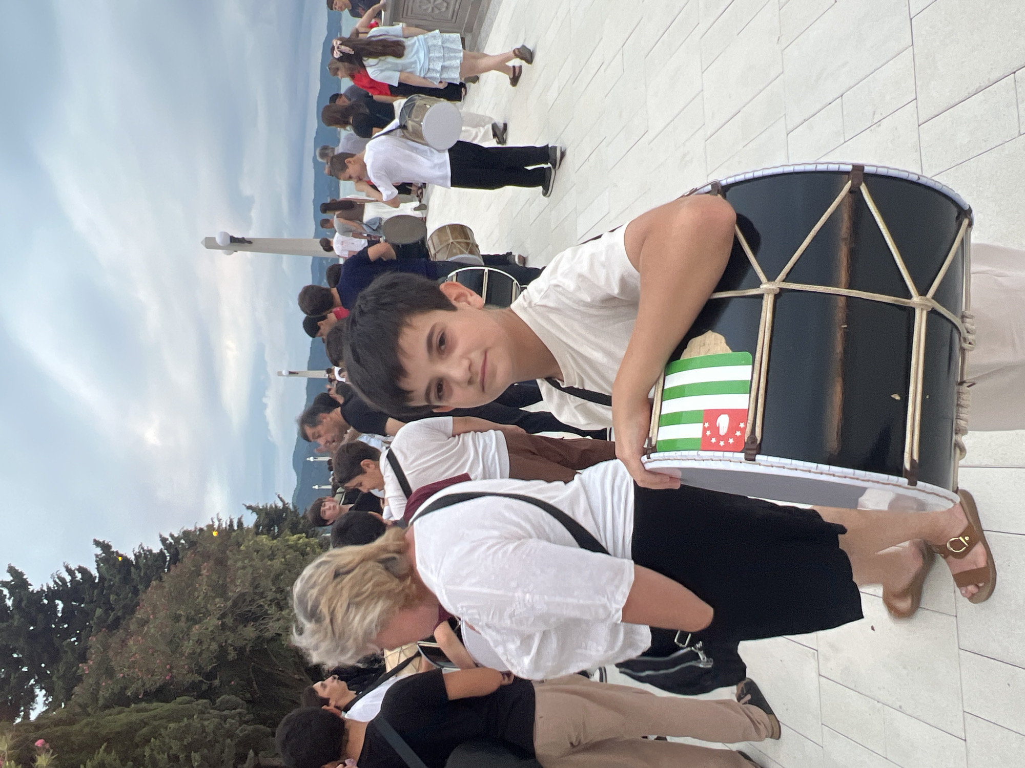 The image captures a daytime scene with a group of people gathered outdoors, possibly for a cultural event or performance, in Sokhumi, Abkhazia. A young boy in the foreground is holding a large drum, which is decorated with green and white stripes and a red star-like emblem. He is wearing a light-colored t-shirt and light-colored pants. To his left, a woman with blonde hair is visible from the back, wearing a white top and a black skirt, with a black bag slung over her shoulder. Other individuals are present in the background, some of whom are also holding drums. The setting appears to be a paved area near a body of water, with trees and distant hills visible. The sky is overcast with clouds.