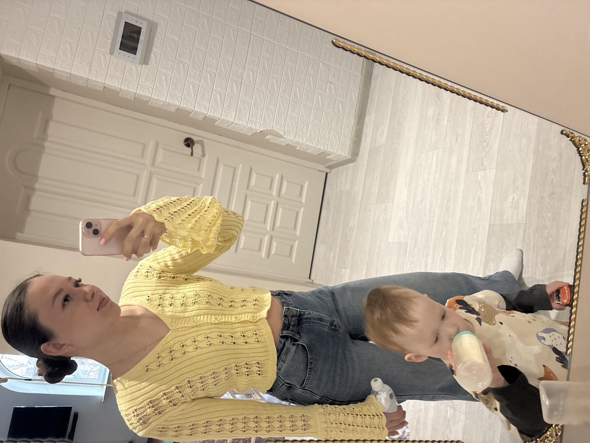 A woman is taking a mirror selfie with a baby/toddler in an indoor setting, likely in Ферма, Russia. The scene features a modern, brightly lit space with a large ornate golden-framed mirror.
The woman, with dark hair pulled back in a bun, is holding a pink smartphone to take the picture. She wears a light yellow, open-knit long-sleeve crop top and blue jeans. A wedding band is visible on her left ring finger, and her nails are painted a light color. Her expression is calm and neutral. She also holds a small clear bottle, possibly hand sanitizer or water, in her left hand.
Standing or being held next to the woman, the baby/toddler has light brown hair and is actively drinking from a baby bottle with a light green cap. They are wearing a patterned long-sleeve top over a dark undershirt and possibly dark pants. In their right hand, the toddler holds a small red and orange toy car.
The background visible in the mirror's reflection includes a white wall covered with textured, rectangular wall panels, resembling bricks or tiles. Below these panels is a white paneled door with a dark doorknob. The flooring is light-colored, resembling wood or laminate planks. A small white electronic device, possibly an intercom or thermostat, is mounted on the textured wall above the door. Natural light suggests it is daytime. No discernible text is visible on any objects.