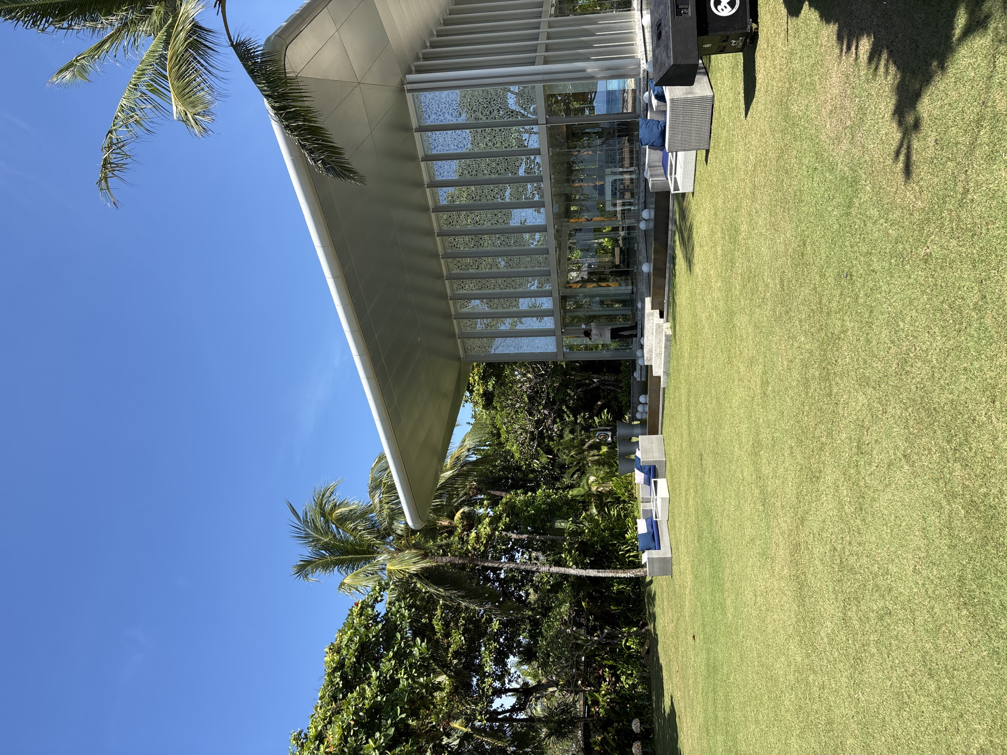 The image shows a modern building with a glass facade in Kuta Selatan, Indonesia, viewed from a slightly low angle. The building has a sharply angled, light-colored roof. The glass facade features a delicate, patterned design. A palm tree is visible on the left, framing the structure. Lush, green foliage surrounds the building. A well-maintained lawn extends to the right. Outdoor seating with blue cushions is arranged near the building. A large speaker sits at the corner of the building. The sky is clear and blue.