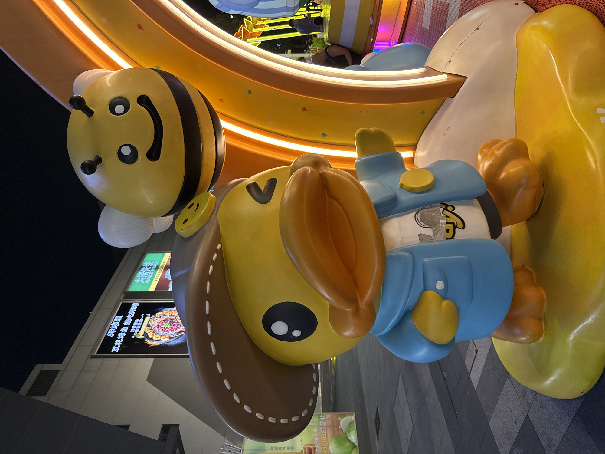 An outdoor nighttime shot captures a large, cartoonish statue of a yellow duck wearing a brown cowboy hat and a blue jacket. The duck has a prominent orange beak and is posed with one hand extended to the right. Above and behind the duck, a smiling cartoon bee with white wings hovers. The statue is situated in front of a brightly lit, curved orange structure with neon lighting. In the background, there are buildings with illuminated signs, including one that appears to advertise a restaurant with a dish of food. The ground is paved with square tiles, and a person is visible in the background near the illuminated structure, appearing to interact with an amusement machine. The overall scene suggests a public space, possibly a shopping area or entertainment district in Tianhe District, China.