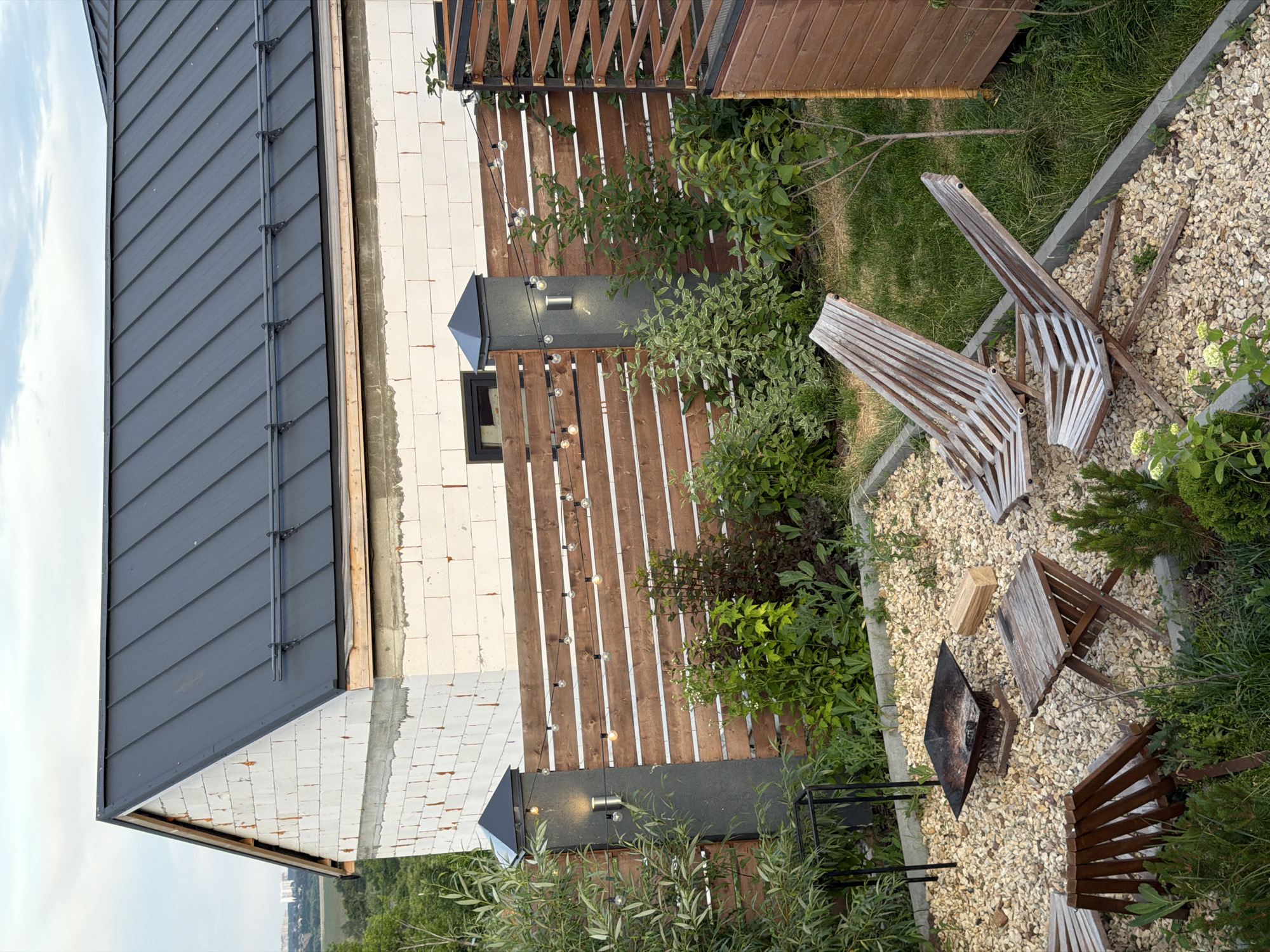 The image depicts a residential backyard in the city of Semiluki, Russia. The scene is dominated by a partially constructed house with white brick walls and a dark metal roof. Adjacent to the house is a wooden slatted fence adorned with string lights. In the foreground, a gravel patio area features two weathered wooden lounge chairs and a small wooden folding table. A square metal fire pit with remnants of burned wood is also present. Surrounding the patio is lush greenery, including bushes, trees, and a manicured lawn. The sky above is overcast.