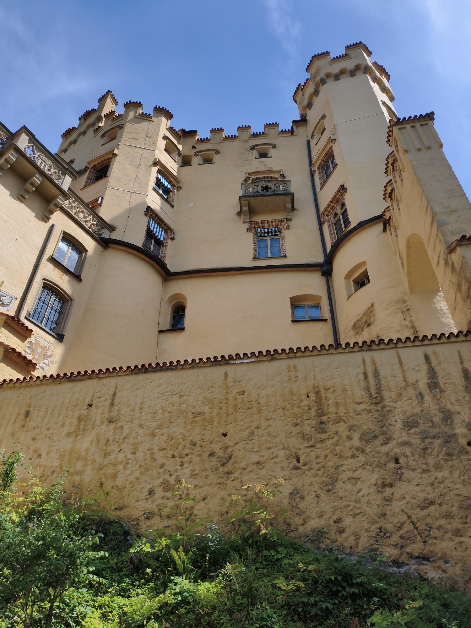 This is a low-angle, upward shot of Hohenschwangau Castle in Schwangau, Germany. The castle is made of pale yellow stucco with a red-tiled roofline and crenellated battlements. Several windows are visible, some with awnings. A stone wall runs along the bottom of the image, overgrown with lush green foliage. The sky is a bright, clear blue. The image captures a sunny day, and there are no people or animals visible.