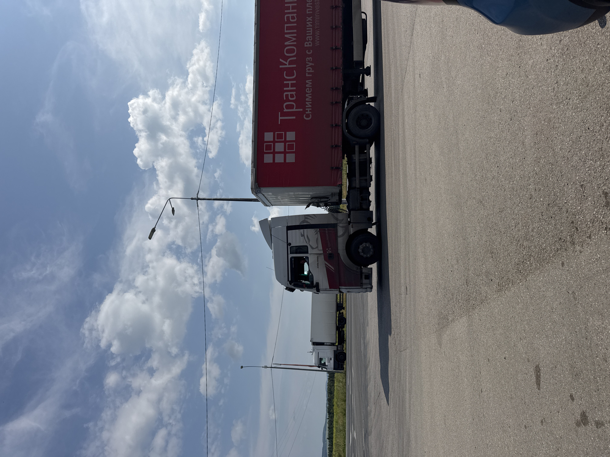 The image depicts a cloudy sky with scattered white clouds over a rural landscape. In the foreground, a paved road is visible, with cracks and some dark stains on its surface. To the right, a portion of a blue vehicle is parked. In the mid-ground, a white semi-trailer truck with a red trailer is parked on the road. The trailer has a logo and text in Russian. Behind this truck, another white semi-trailer truck is parked. A light pole stands between the two trucks. The background shows a green field and a line of trees in the distance, suggesting a location outside of a dense urban area. The overall scene appears to be a roadside area, possibly a parking or rest stop. Based on the provided context, the location is Verkh-Sosnova, Russia.