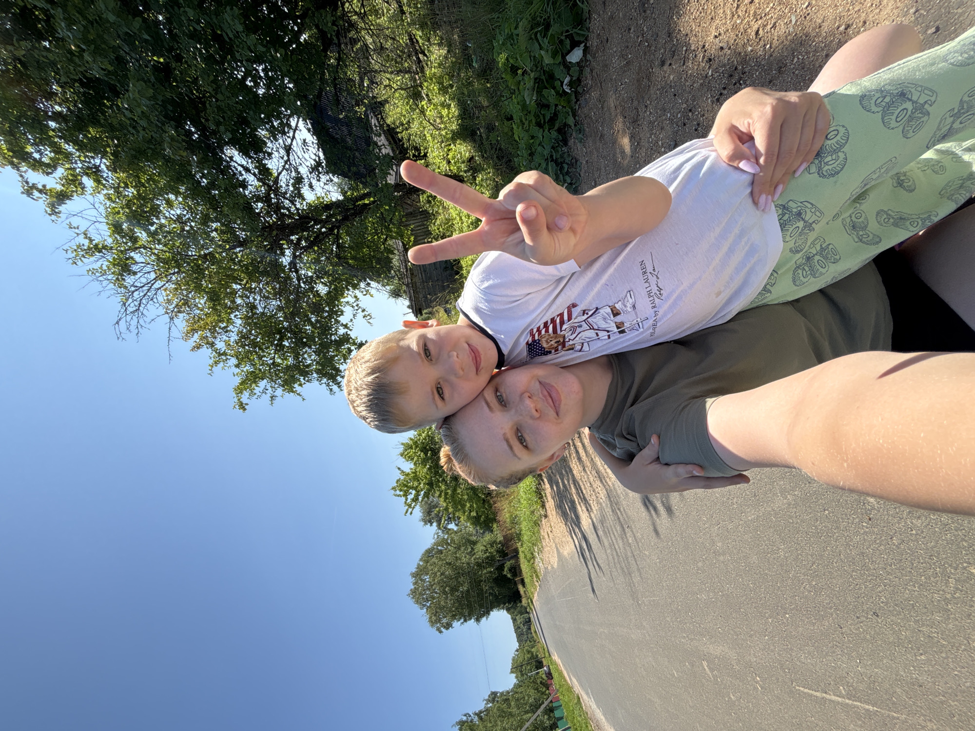 A mother and her young son are taking a selfie outdoors on a sunny day. The mother is in the foreground, wearing a green t-shirt and light green pants with a monster truck pattern. Her arm is around her son, who is positioned above her. The son is wearing a white t-shirt with a graphic design on it. He is making a peace sign with his left hand. The background shows a road, trees, and a clear blue sky. The image appears to be taken in the afternoon due to the sunlight and shadows. The overall mood is happy and relaxed. Text on the son's shirt reads: "POLO G.E.A. BY RALPH LAUREN". The scene is consistent with a rural or suburban setting in Дунилово, Russia.
