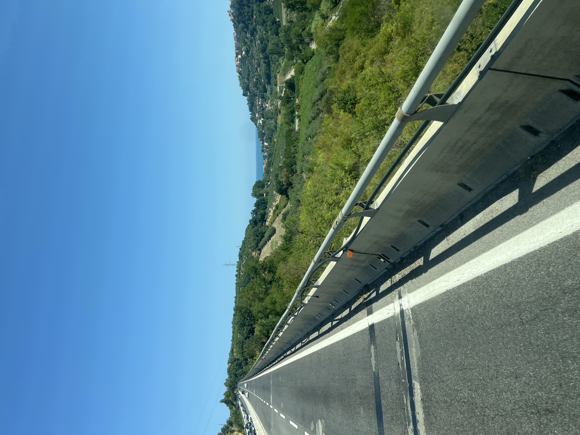 An elevated highway curves to the left under a clear blue sky. The highway is asphalt with white lane markings and a concrete barrier with a metal railing runs along the right side. The surrounding landscape is hilly and green, with trees and olive groves. In the distance, a coastal town with buildings is visible on a hill overlooking the sea. There are no people or animals in the image. The perspective suggests the photo was taken from a moving vehicle.