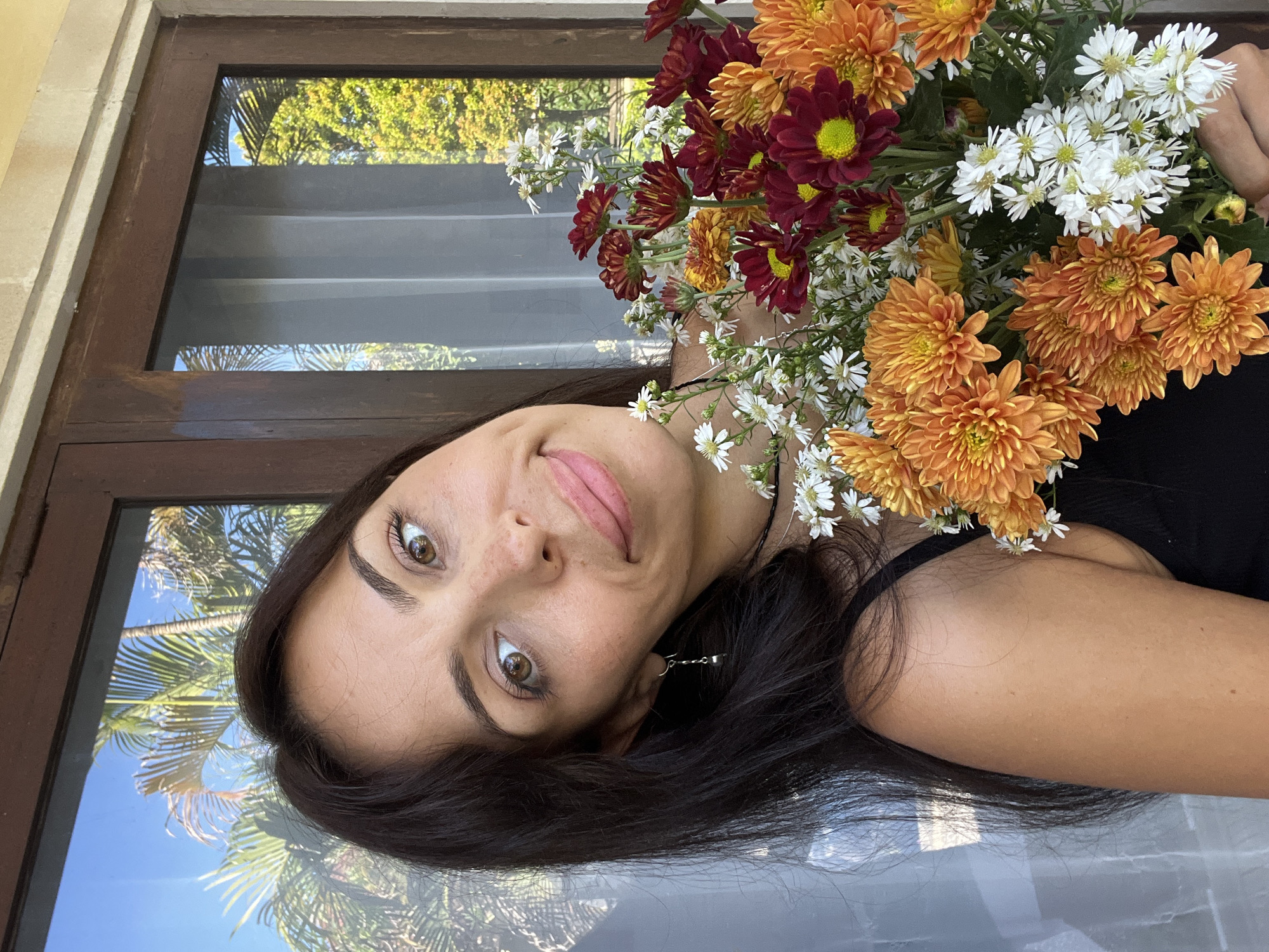 The image presents a casual portrait of a woman lying down, with her head positioned towards the left side of the frame, looking upwards and directly at the viewer. She has long, dark hair spread out, and her skin tone is medium with visible freckles across her nose and cheeks. Her eyes are light brown, and she has a gentle, slightly amused smile on her lips, which are adorned with light pink lipstick. A small, silver-toned earring is visible on her left ear, but any specific design or text on it is indistinct. She is wearing a dark-colored, possibly black, top.
A large, vibrant bouquet of flowers rests on her right side, partially covering her shoulder and upper chest. The bouquet comprises numerous orange chrysanthemums, several dark red (maroon) chrysanthemums with prominent yellow-green centers, and delicate clusters of small white daisy-like flowers.
The background features a dark brown wooden window frame with glass panes. Reflected prominently in the glass are lush green tropical foliage, including discernible palm fronds, set against a bright blue sky, suggesting a sunny outdoor setting. Faintly visible through the glass, behind the reflections, are horizontal gray slats, possibly internal blinds or shutters. The surface beneath the woman, visible towards the bottom of the frame, is reflective, appearing to be a light-colored, possibly polished, floor or surface, also showing subtle reflections. The bright lighting, tropical plants, and clear sky indicate it is daytime, capturing a pleasant and serene moment.