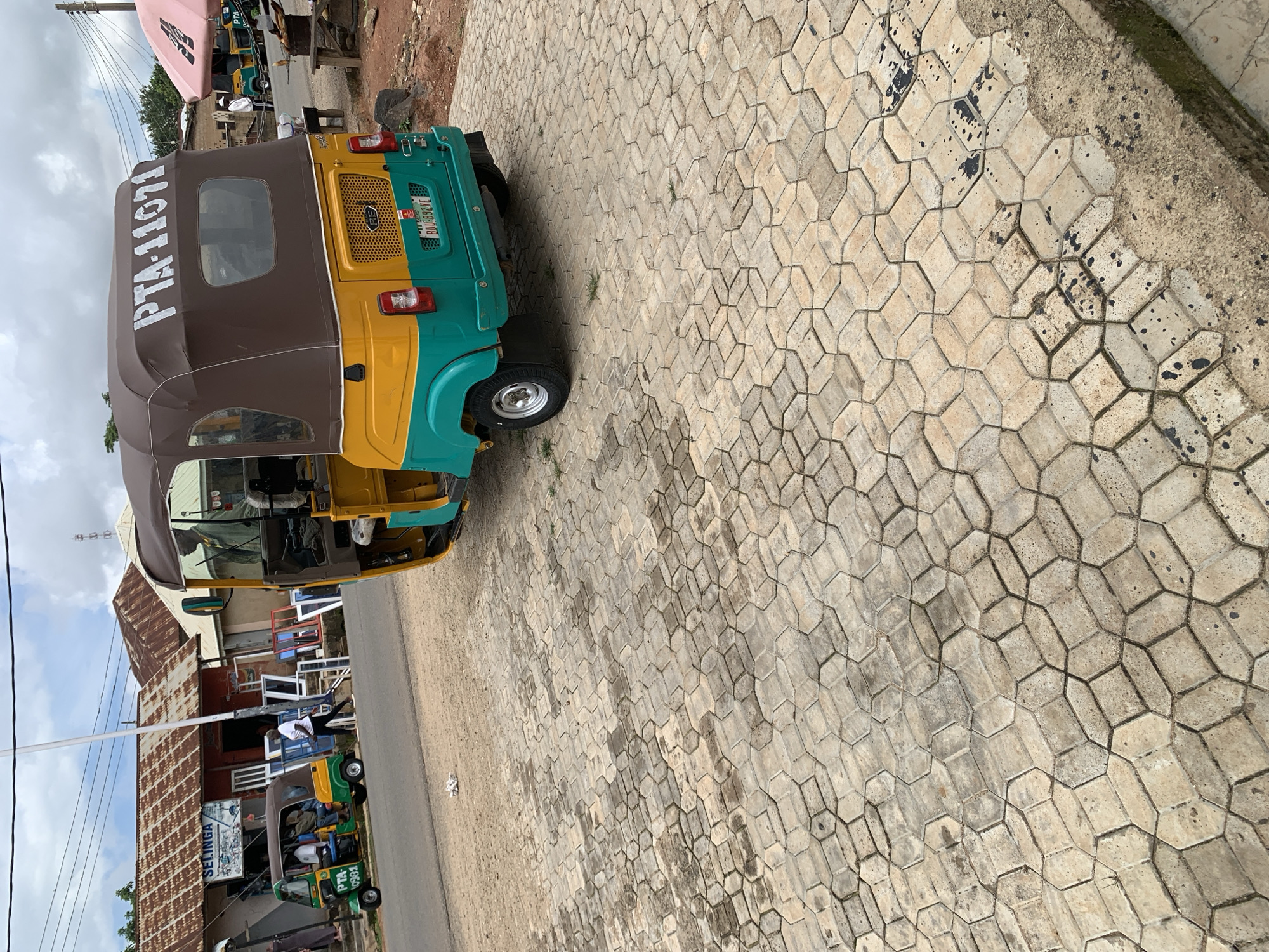A yellow and teal auto rickshaw, identified by the license plate PTA-11071, is parked on a paved surface. The auto rickshaw has a brown canopy and is positioned on the right side of the image, facing away from the viewer. In the background, a green auto rickshaw with the license plate PTA-0991 is visible, carrying passengers. Several buildings with corrugated tin roofs line the street, and windows are displayed outside one of the establishments. A person in a white shirt and dark pants walks in the middle ground, while another individual in a green jacket is seated in the auto rickshaw in the foreground. The environment appears to be a commercial area with overhead wires and a cloudy sky.