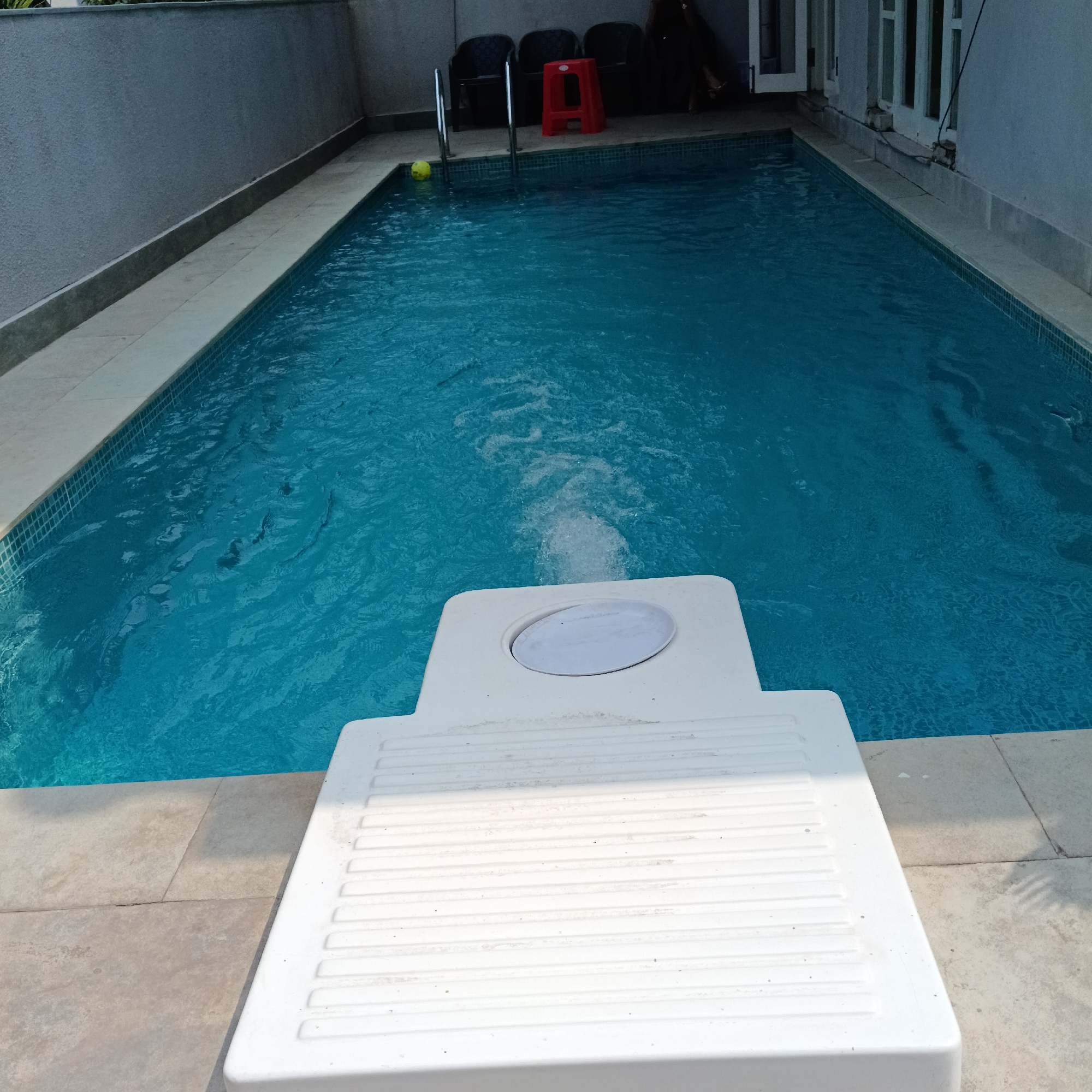 The image depicts a rectangular swimming pool, filled with blue water, located in a partially enclosed space. A white, rectangular diving board or platform is situated at the edge of the pool in the foreground. Behind the pool, there's a row of black plastic chairs and a red plastic stool next to a metal ladder. The pool is bordered by tiled surfaces. The background includes a light grey wall on one side and a building structure with white-framed windows and doors on the other. A yellow ball floats near the ladder. The location is speculated to be Calangute, India. No people are visible in the immediate scene but a pair of bare feet and legs are visible behind the chairs.