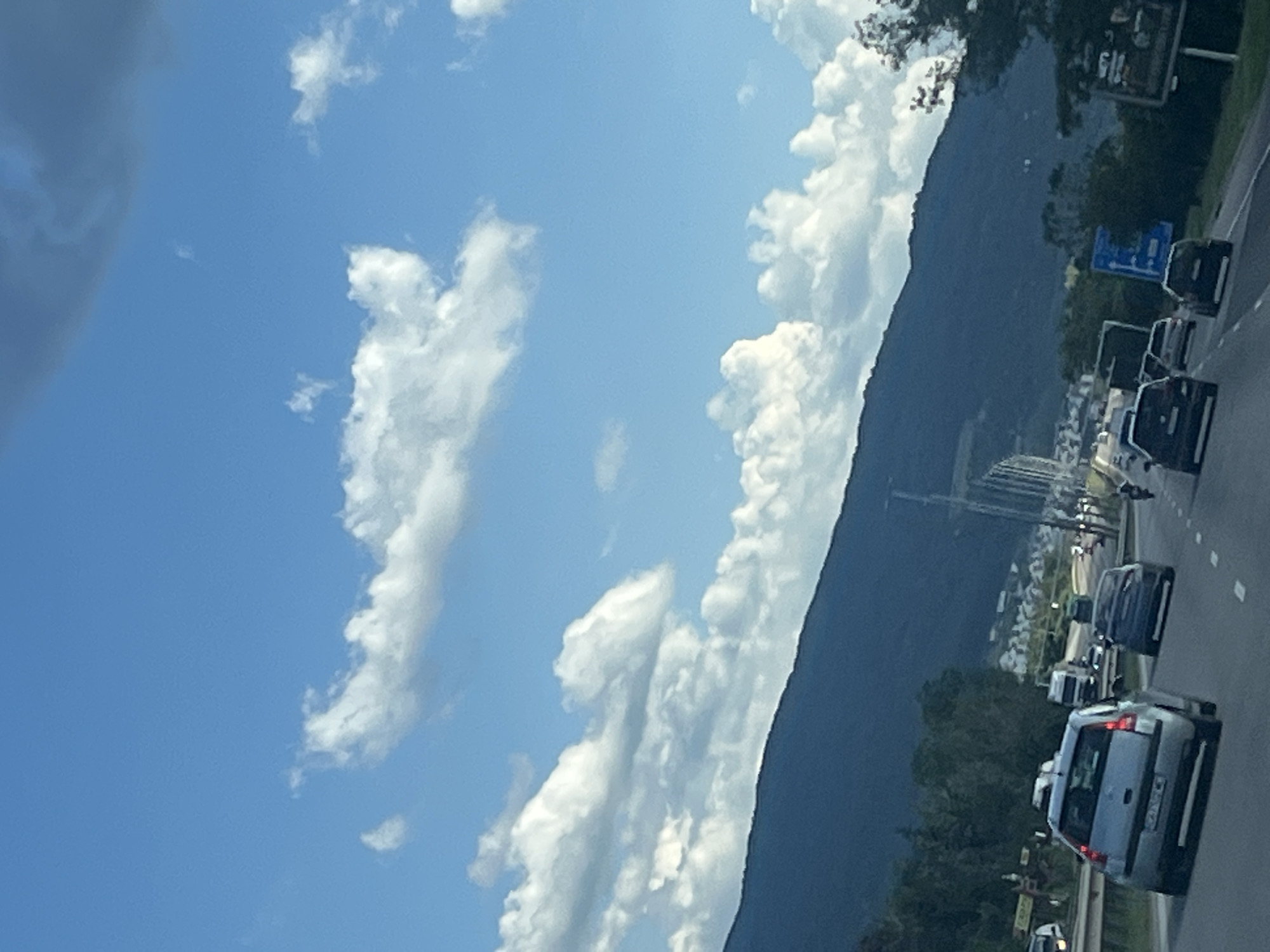 The image depicts a highway scene with vehicles moving along multiple lanes. In the foreground, a silver hatchback is visible, followed by several other cars and a truck. The highway is bordered by a guardrail on the left and trees and a dark, mountainous landscape in the background. The sky is blue with scattered white clouds, and sunlight suggests it is daytime. There are buildings visible on the slopes of the mountain.