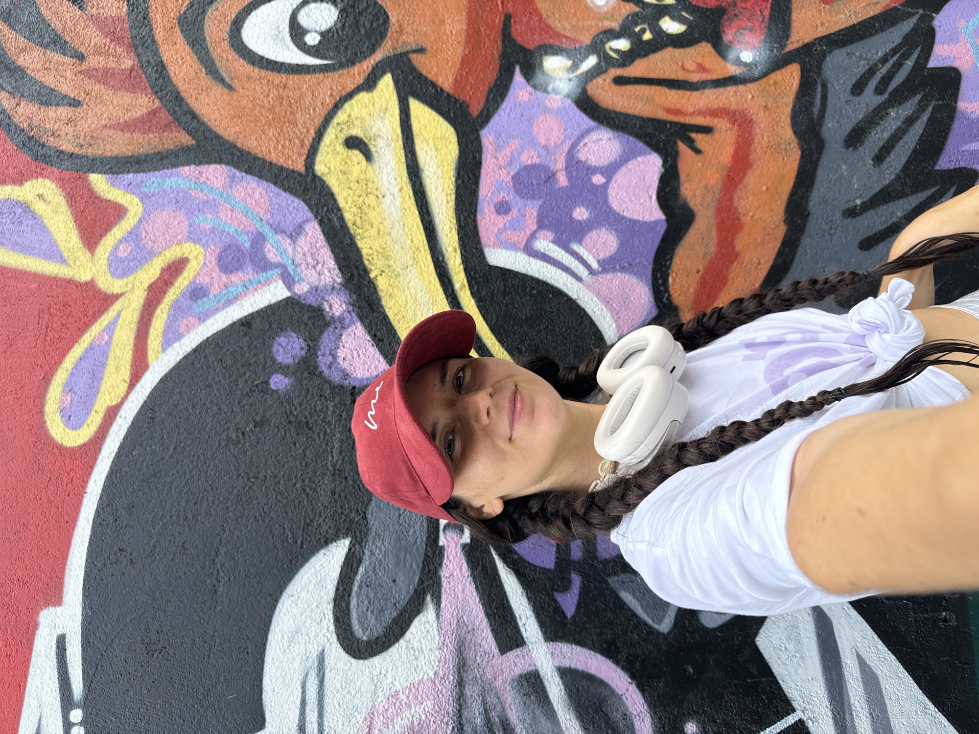 A young woman with dark hair braided into pigtails and a red baseball cap stands in front of a graffiti-covered wall. She is wearing a white t-shirt and white headphones around her neck. The graffiti features colorful abstract shapes and a cartoonish orange bird with a yellow beak and large eyes. The image appears to be a selfie, taken from a slightly low angle. The location is Phu Thuy Ward, Vietnam.