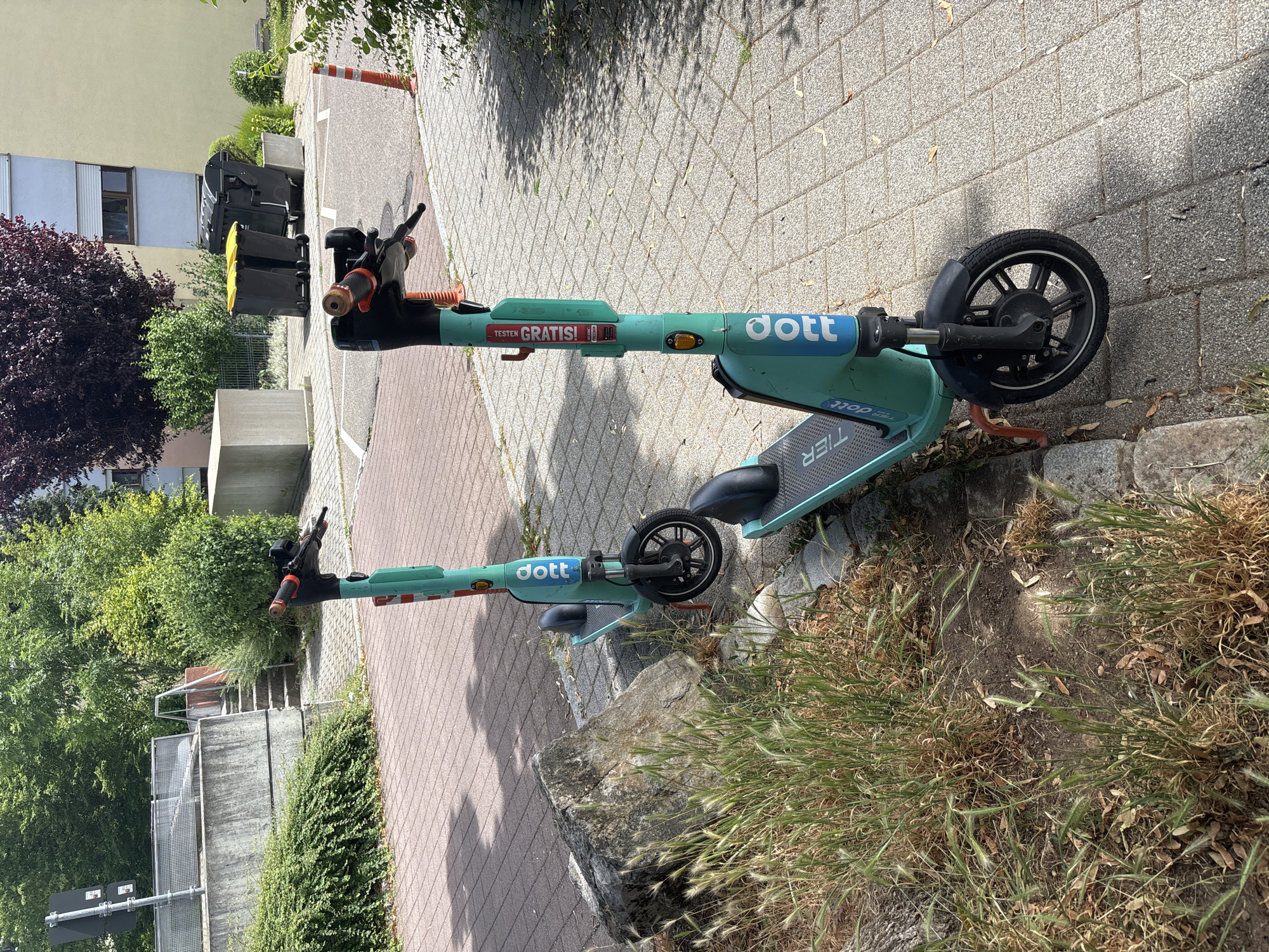 Two teal colored electric scooters are parked on a brick walkway in front of a grassy area with a large rock. The scooters are positioned side-by-side, facing forward. The scooter on the right has "dott" printed in white lettering on its side. The scooter on the left also has "dott" printed on its side. In the background, there are buildings, trees, and garbage bins. The environment is outdoors on a sunny day.
