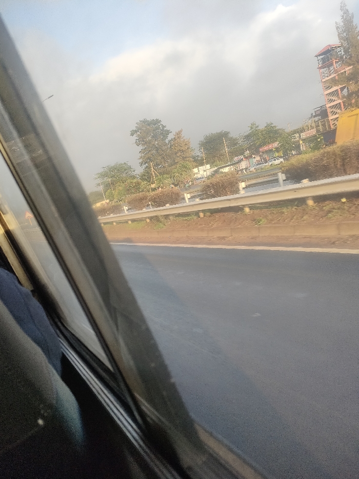 The image depicts an oblique exterior view captured from within a vehicle, showing a paved road and the surrounding environment in Theta ward, Kenya.
In the immediate foreground on the left, elements of the vehicle's interior are visible, including a dark window frame and dark fabric, possibly a seat or clothing. The primary subject is a multi-lane asphalt roadway, dark grey in color, extending from the midground into the distance. A solid white line delineates the right edge of the visible road segment.
Parallel to the road, a white metal guardrail is present, separating the road from an earthen strip. Beyond this strip, a row of low, bushy green and brown shrubs forms a median or roadside barrier. In the background, numerous trees with varied foliage densities and heights line what appears to be another roadway or area with commercial structures. Distant buildings are partially obscured by the trees.
To the far right of the frame, a distinctive, multi-story structure with an exposed, reddish-brown skeletal frame, possibly a tower, stands prominently against the skyline. Several vehicles, including at least one white car, are discernible on a road or parking area beyond the shrubbery. The sky above is partly cloudy, showing patches of light blue and scattered white-grey clouds, indicating daylight conditions. No individuals are clearly visible.