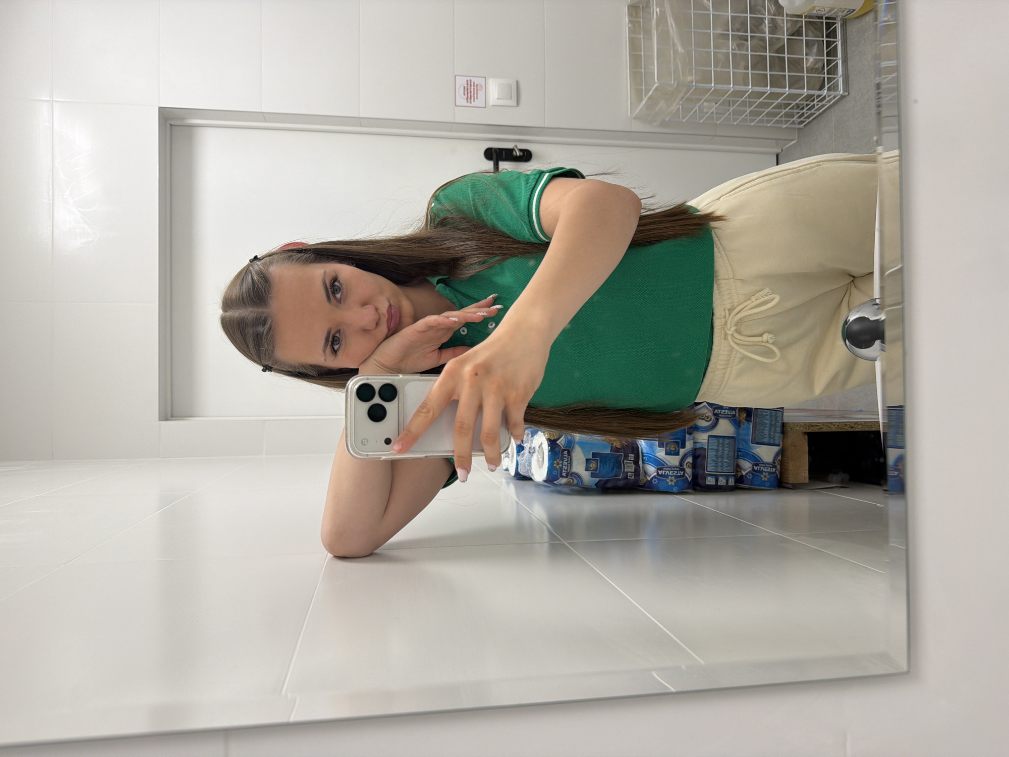 A young woman is taking a selfie in what appears to be a bathroom. She is wearing a green polo shirt with white trim and light-colored sweatpants with drawstrings. Her dark brown hair is styled with clips. She is holding a smartphone up to take the picture, resting her head on her hand. Behind her, stacked under what seems to be a sink or shelf, are packages of blue and white toilet paper. The bathroom has white walls and white tiled floors. A white towel rack or shelf is visible above her head. A white door with silver hardware is also visible in the background. Based on the packaging of the toilet paper, it may be possible to determine the precise geographic location of the setting in the city of Sofia, Bulgaria. No specific actions, interactions, or other people are visible in the frame.