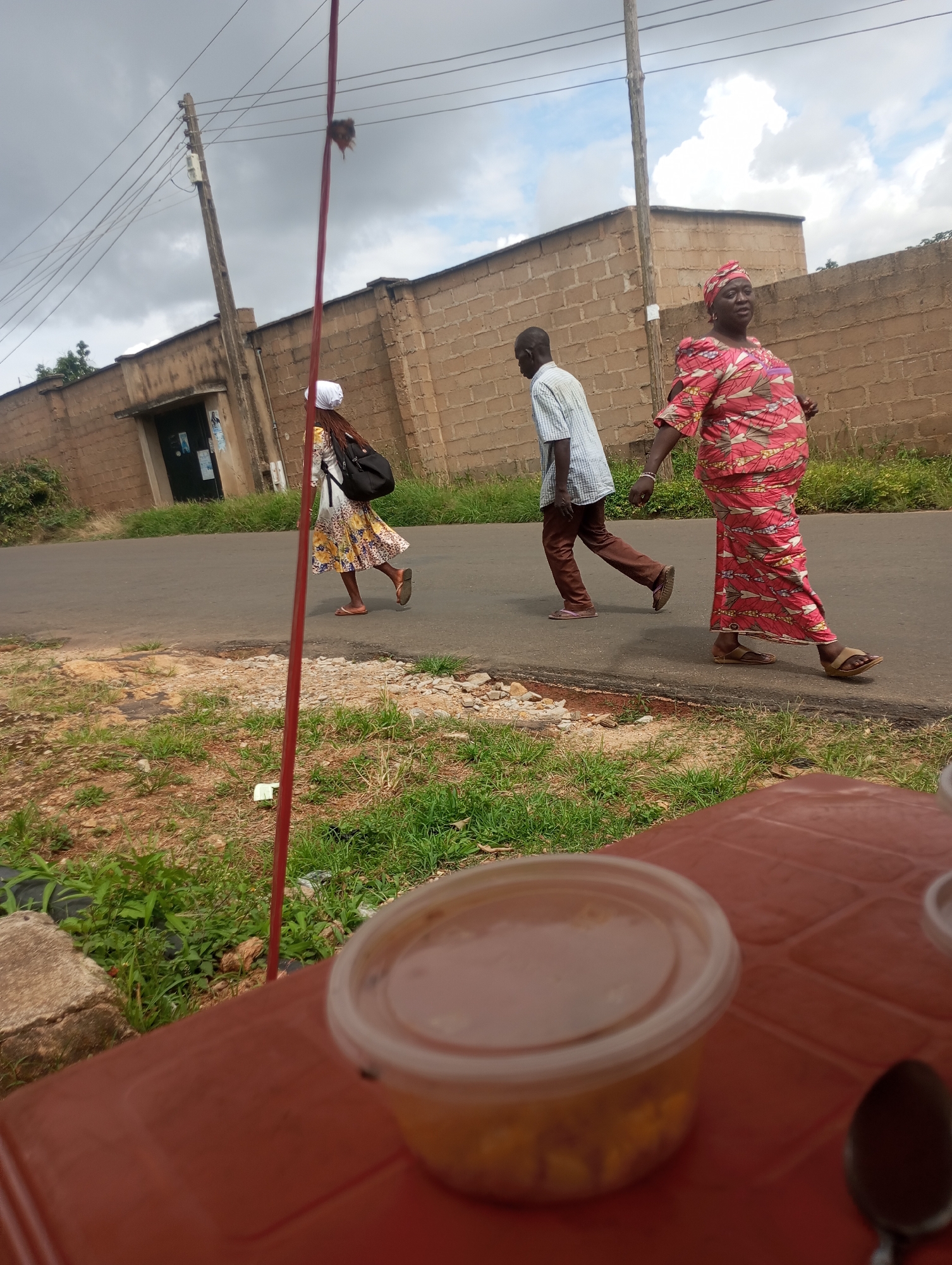 The image depicts a street scene in Jos, Nigeria, under an overcast sky. In the foreground, a red plastic table holds a clear plastic container of food and a spoon. The table is positioned on the right side of the frame. The background reveals a paved road, with a woman in a vibrant red and yellow patterned dress and headscarf walking towards the right. She is followed by a man in a striped blue and white shirt and brown trousers, also walking to the right. Further to the left, a young woman in a white headscarf and a patterned yellow and white dress with a black backpack is walking away from the viewer, also towards the right. Behind them, a brick wall with a building facade is visible, and utility poles with electrical wires traverse the sky. The ground on the left side of the road is covered in dry grass and some stones.