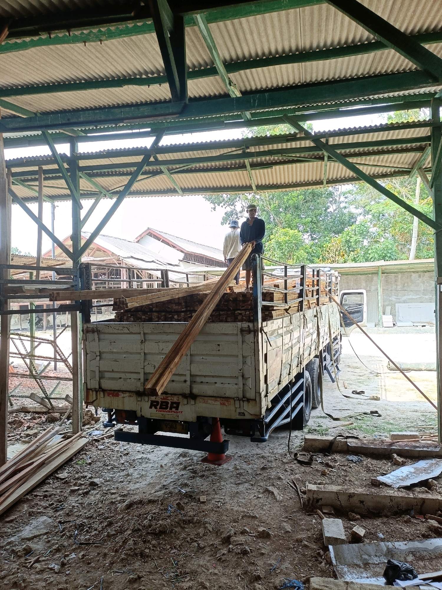The image depicts an outdoor scene within what appears to be a lumber yard or workshop in Pordapor, Indonesia. In the foreground, a flatbed truck is laden with stacks of lumber. Two men are visible on top of the truck, one standing and holding a long piece of wood, appearing to be engaged in the process of loading or unloading. The truck's tailgate is down, and other pieces of lumber are also scattered on the ground around it. The environment shows signs of construction or renovation, with debris and unfinished structures visible. The structure overhead, with its corrugated roof and exposed beams, suggests an open-air workspace or storage area. In the background, trees and other buildings indicate a semi-rural or industrial setting. The lighting suggests it is daytime, possibly late morning or early afternoon.