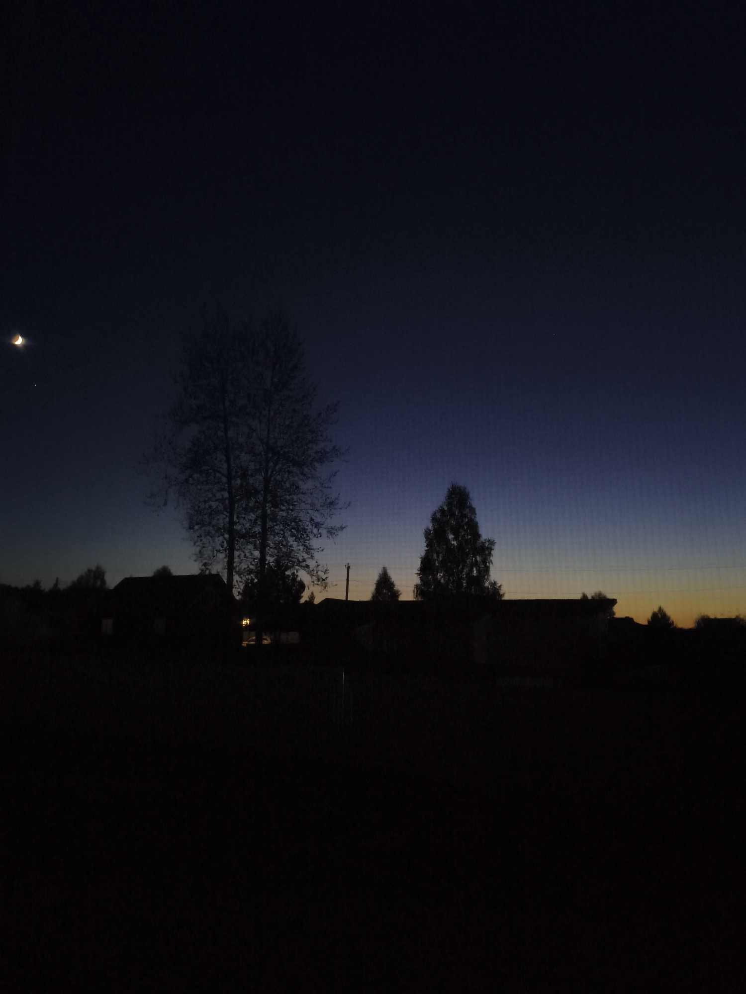 The image shows a nighttime scene in Русский Вожой, Russia. A crescent moon is visible in the upper left of the image. Silhouetted houses and trees are visible against a dark blue and orange gradient sky. The foreground is completely dark. A single light is visible in the distance.