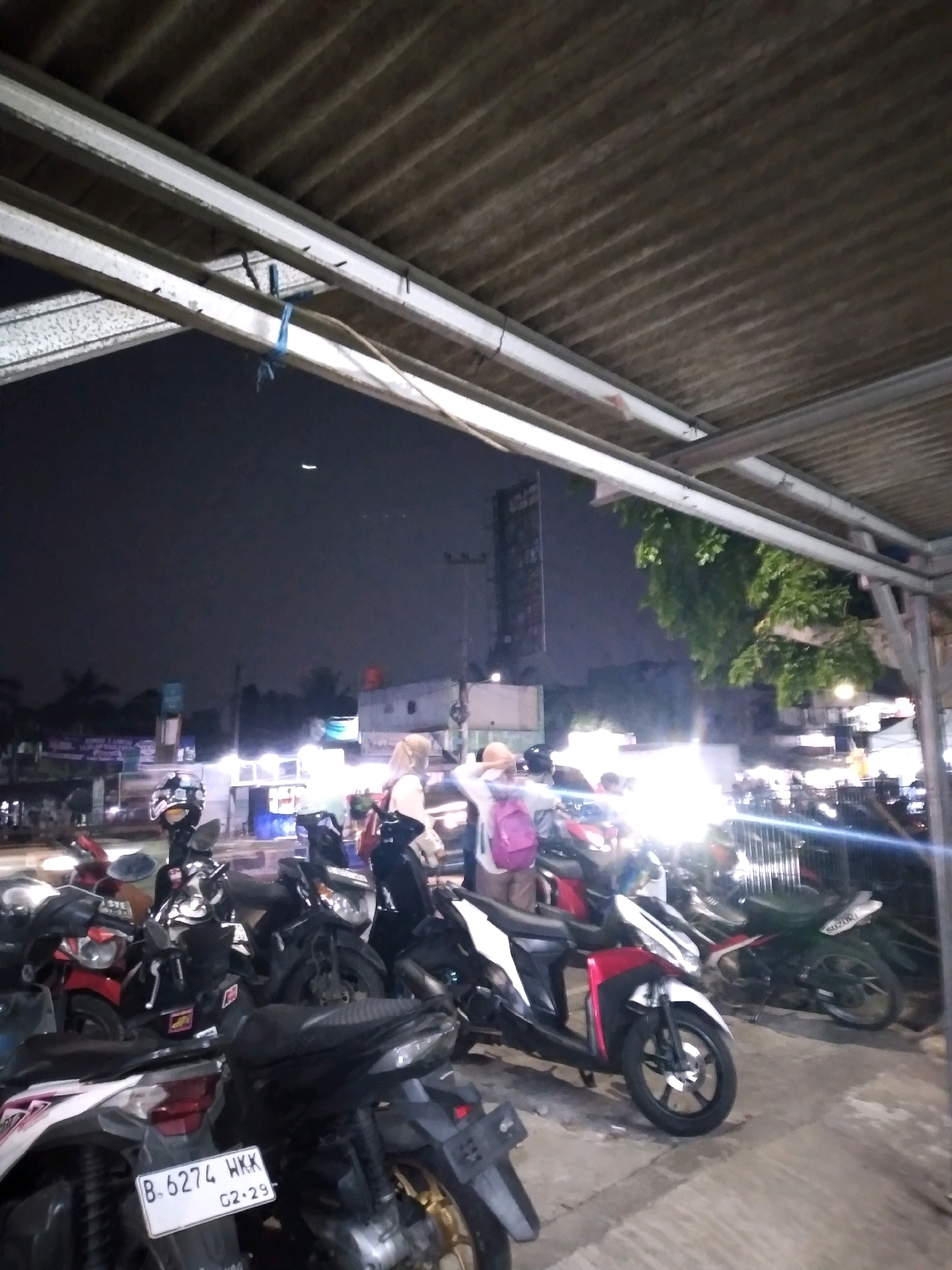 This image depicts a nighttime scene in an urban environment, likely a parking area or roadside stall. Several motorcycles are parked close together, some with their headlights on, illuminating the surroundings. In the background, there are buildings, streetlights, and a dark sky with a crescent moon visible. A few people are present, some standing near the motorcycles, and one is seated. The overall atmosphere is casual and suggests a gathering or a place where people stop. The foreground is dominated by the motorcycles, with the roof structure of a covered area visible at the top. The license plate on one of the motorcycles reads "B. 6274 WKK 02.29".