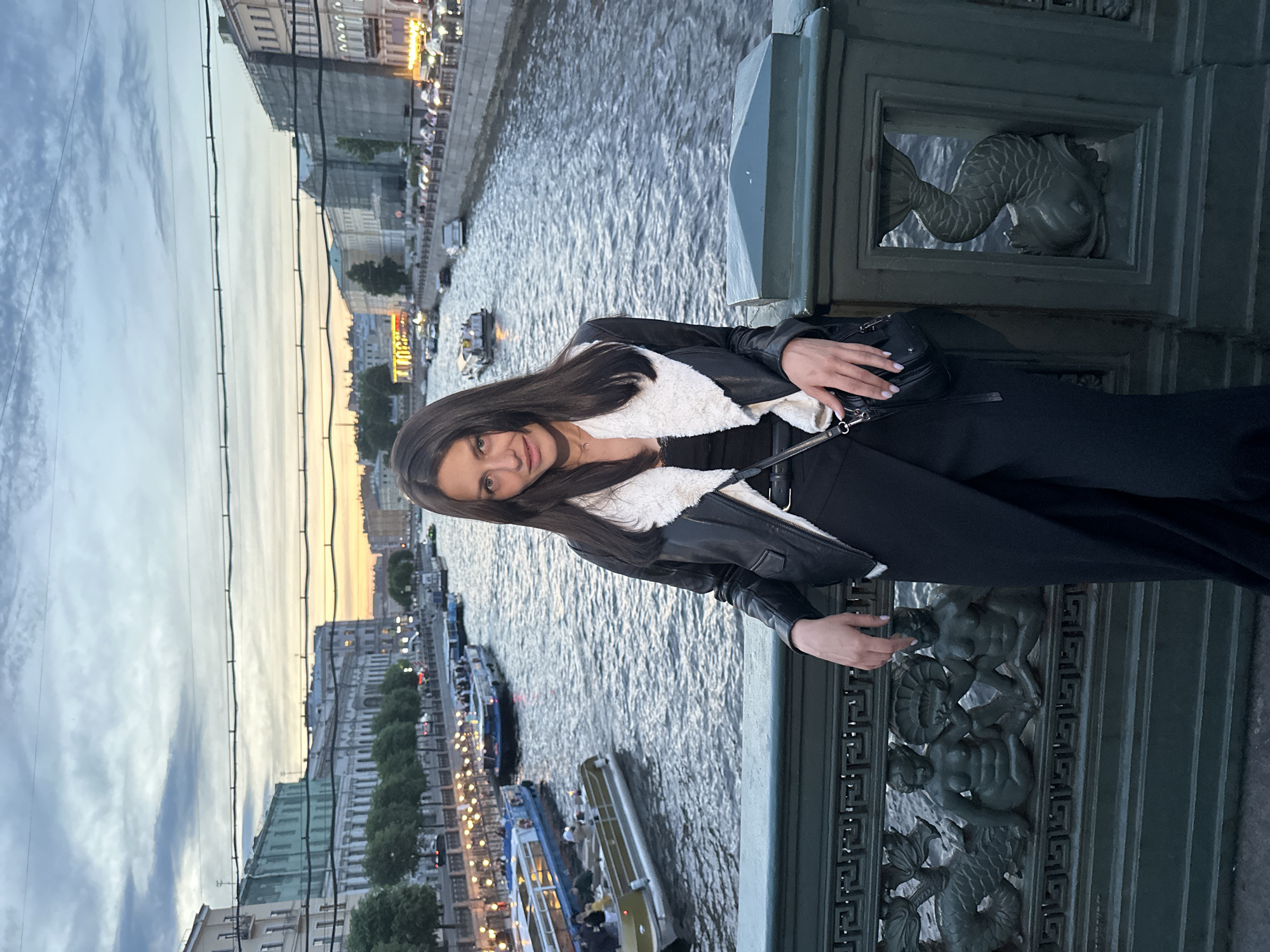 A woman stands on a bridge in Saint Petersburg, Russia, posing for a photo. She is wearing a black leather jacket with a white furry collar, a black top, and black pants. She has a black purse slung over her shoulder. The bridge has ornate dark green metal railings with decorative figures of mermaids and fish. In the background, a wide canal or river is visible, with several tour boats and buildings lining the embankments. The sky is overcast with hints of sunset colors.