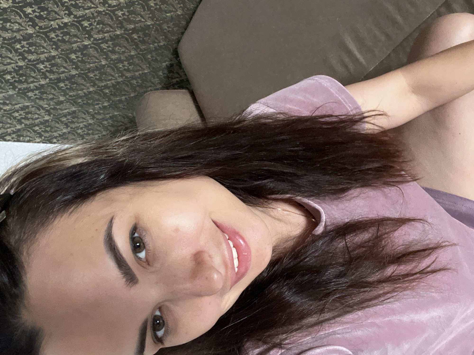 The image is a medium shot selfie of a woman with long, dark hair. She is smiling and looking at the camera. She is wearing a pink t-shirt. The background appears to be a wall with patterned wallpaper and a portion of a couch. The lighting is natural and soft. The overall impression is casual and relaxed.