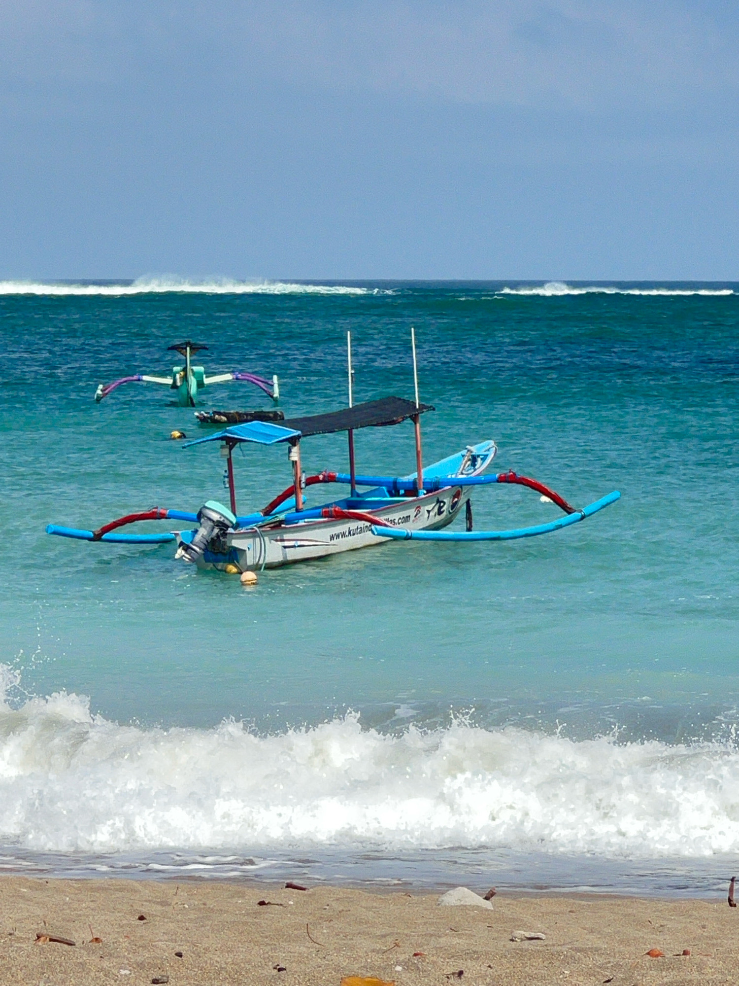 This image depicts a view of the ocean and beach in Kuta, Indonesia. The scene features two traditional Indonesian outrigger boats floating on the turquoise water. The foreground shows the sandy beach with scattered debris. In the middle ground, one boat with a blue canopy and an outboard motor is closer to the shore, with the website "www.kutaind.com" visible on its side. Another outrigger boat is positioned further out in the water. The background shows waves breaking on the horizon under a clear blue sky. The overall impression is a serene tropical beach scene.