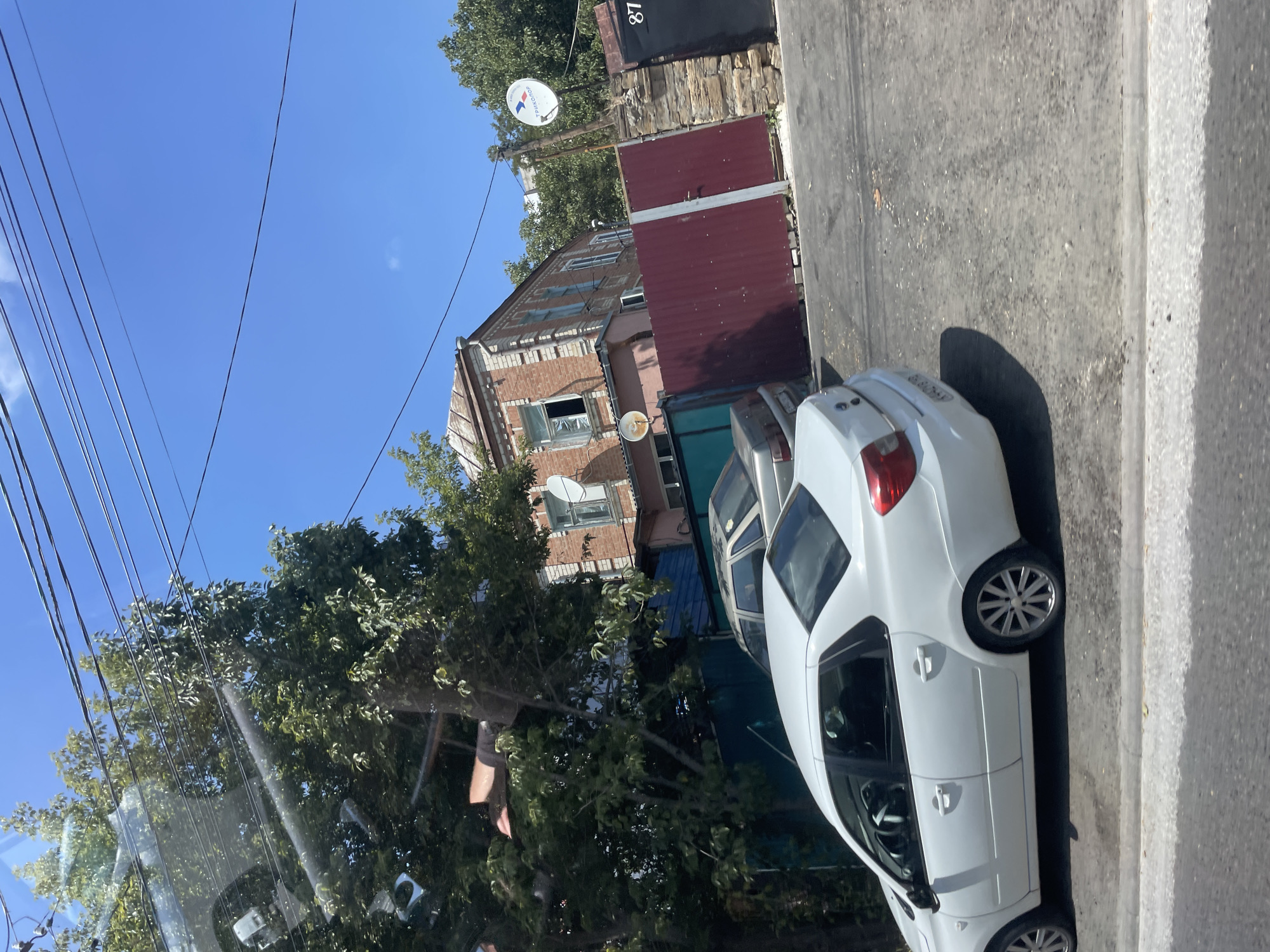 A daytime, eye-level, outdoor shot captures a residential street in Stavropol, Russia, under a clear blue sky. In the foreground, a white sedan is parked, facing right, with its rear portion cropped out of view. Behind it, a silver sedan is partially visible, parked further back. A dark green fence runs along the left side of the image, partially obscured by lush green trees.
The background features a two-story brick building with a light-colored roof and visible windows. A large, dark red metal gate dominates the center of the background, with a stone wall to its right. A satellite dish, bearing the "Tricolor TV" logo, is mounted on a pole near the gate. The number "87" is stenciled in white on a black surface to the far right of the gate. Overhead, a network of black power lines stretches across the frame. The overall scene depicts a typical urban residential area on a sunny day.