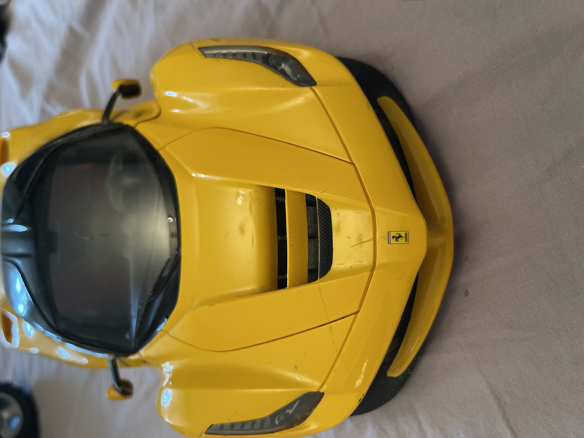 The image shows a yellow toy car, specifically a Ferrari, from an overhead perspective. The car is positioned on a light-colored, textured surface resembling fabric. The car exhibits visible details such as a black windshield, front headlights, a Ferrari logo on the hood, and side mirrors. The scene is indoors, but there is no specific evidence indicating that this picture was taken in Moscow, Russia.