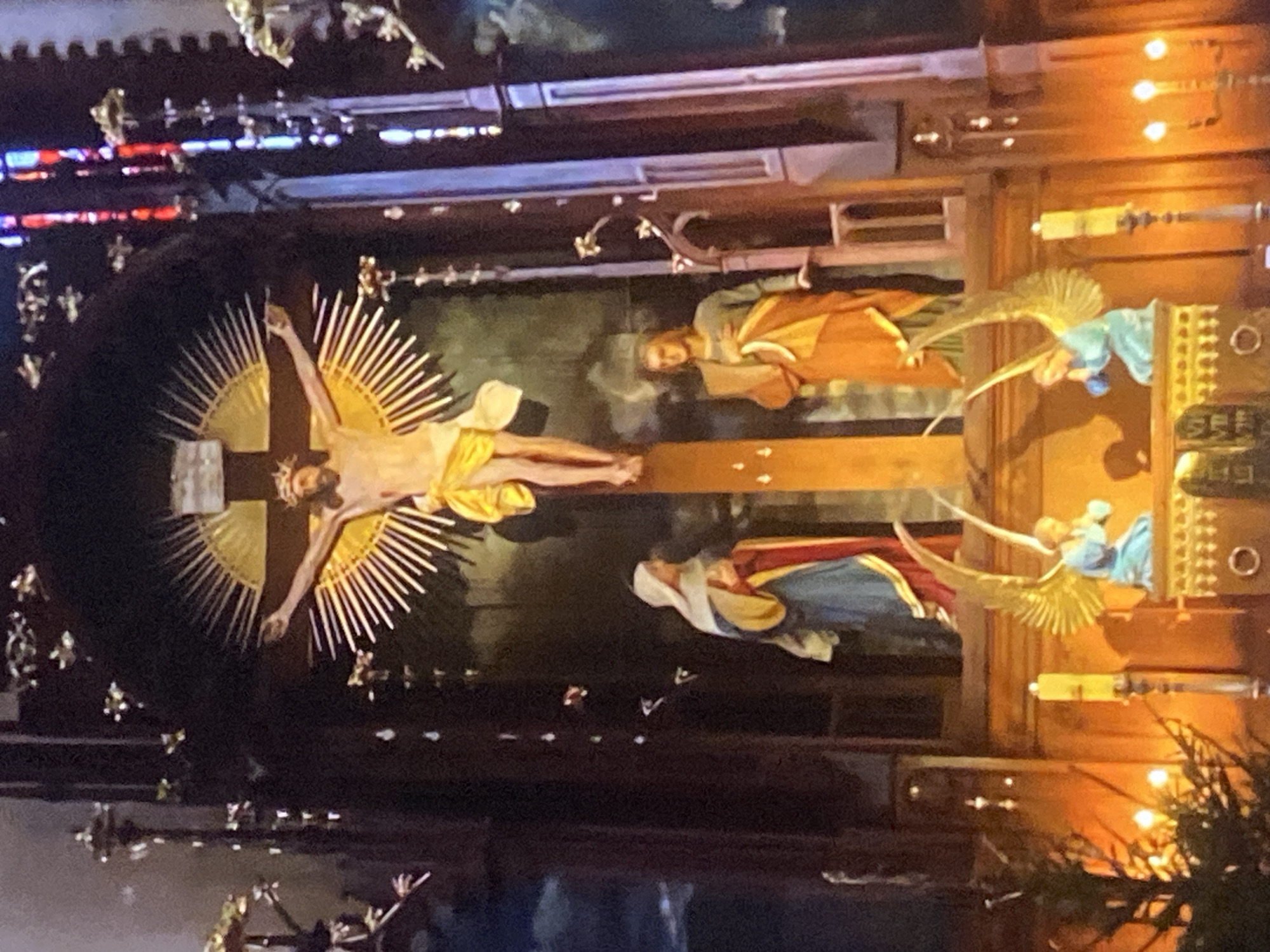 This is a photograph of a religious artwork, likely an altarpiece or diorama, located in Tinaquillo, Venezuela. The central figure is a depiction of Jesus Christ on the cross, surrounded by a radiating halo of golden spikes. The scene includes figures of angels and what appears to be Mary, mother of Jesus, positioned around the cross. The artwork is set against a backdrop of dark tones, with the figures and central cross illuminated. The structure is decorated with intricate carvings and lights, appearing to be part of the interior of a church or religious setting. The presence of decorative elements in the foreground suggests that the photo was taken from the perspective of a viewer inside this religious space.