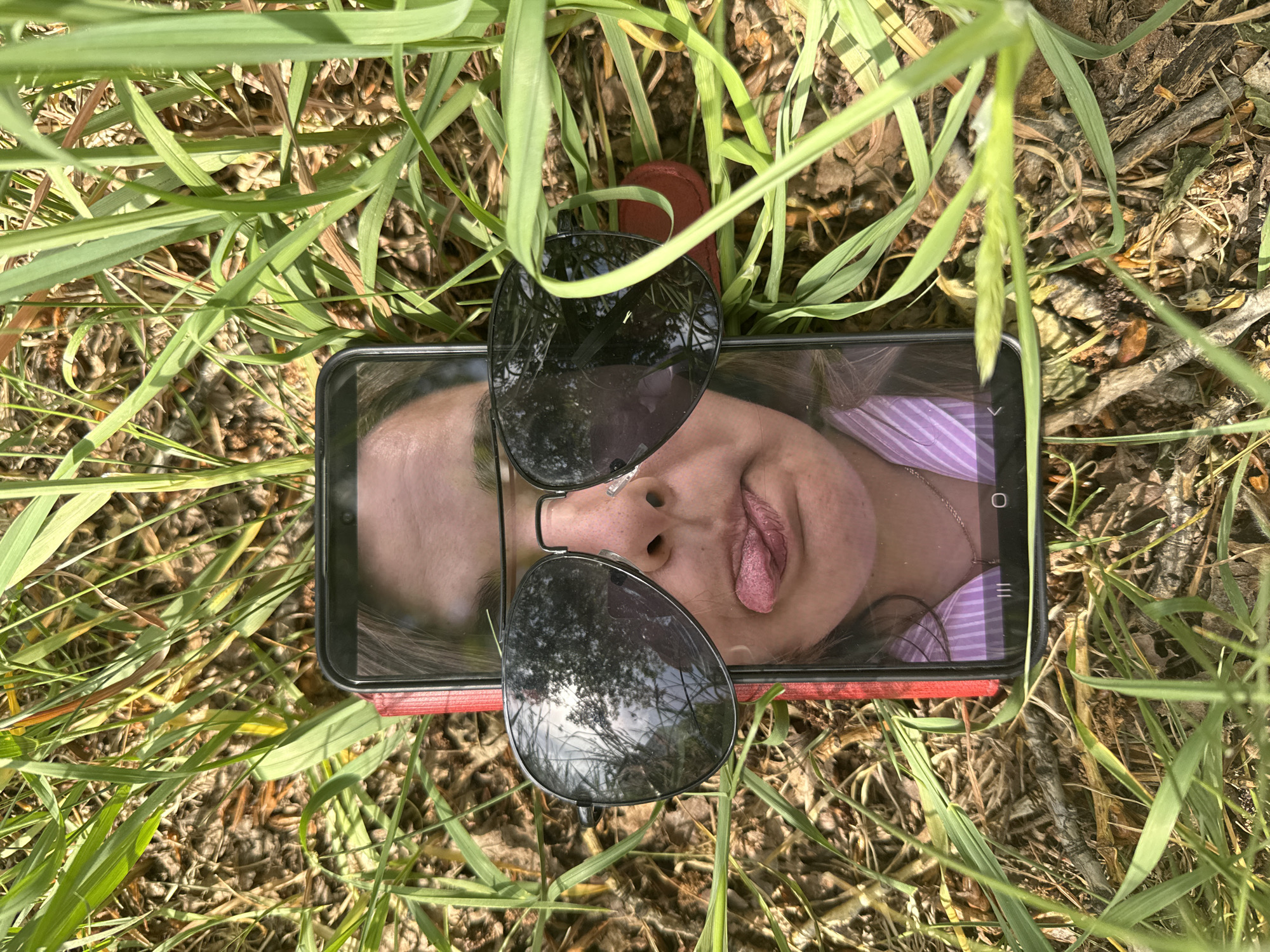 A smartphone rests among blades of green grass and dry leaves, displaying a close-up image of a person with their tongue out. Sunglasses are placed on top of the phone's screen. A red object is positioned beneath the phone. The setting appears to be outdoors in a grassy area. The city of Chaikovsky, Russia is not discernible.