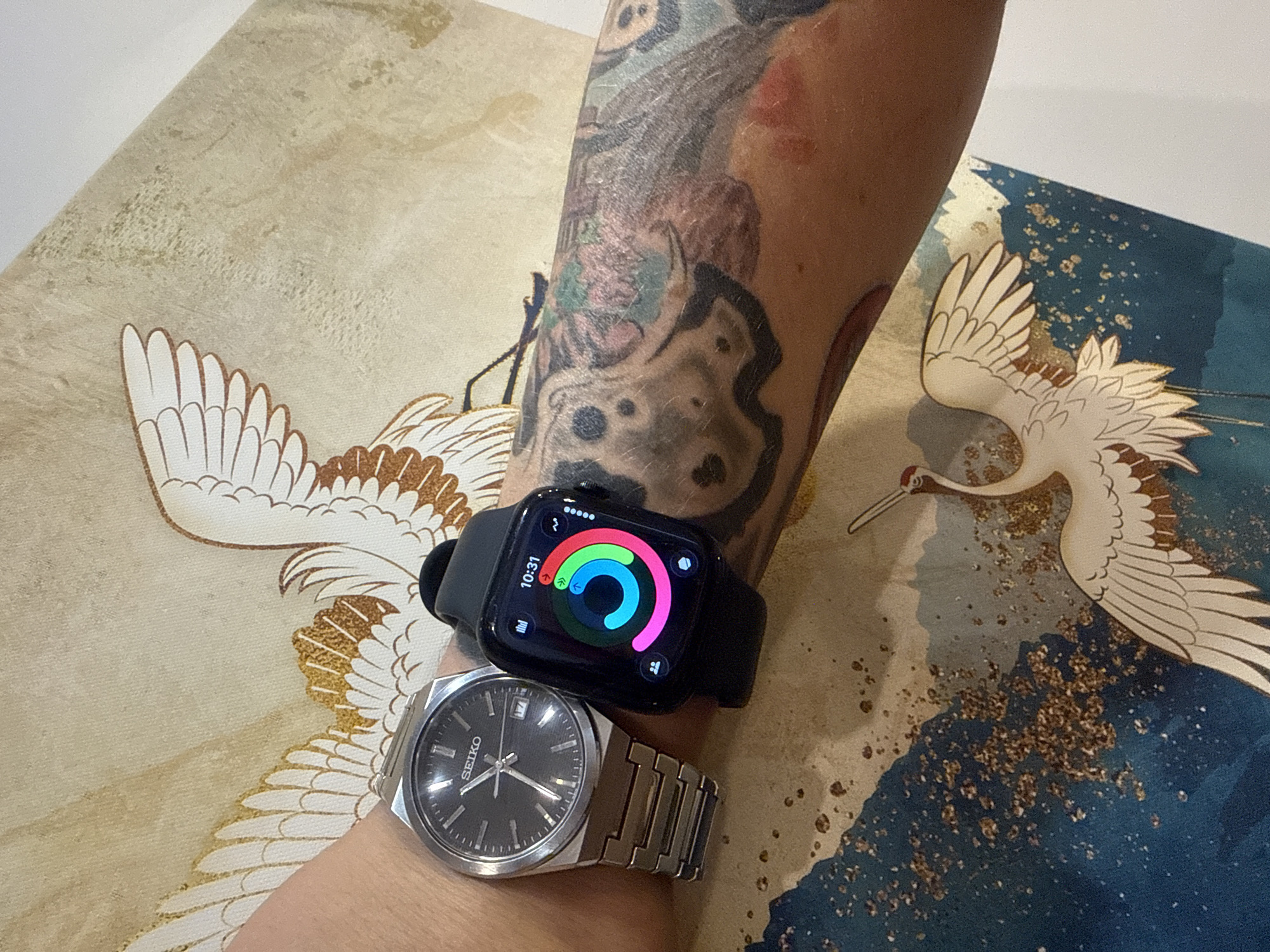This image depicts a close-up view of a person's arm, adorned with tattoos, wearing two wristwatches. One is a silver analog Seiko watch, and the other is a black smartwatch displaying a colorful circular activity tracking interface and the time "10:31". The arm is resting on a fabric background featuring a traditional Japanese art style, with depictions of cranes and gold leaf accents. The scene does not provide any visual information that could indicate a specific location within Vladivostok, Russia.