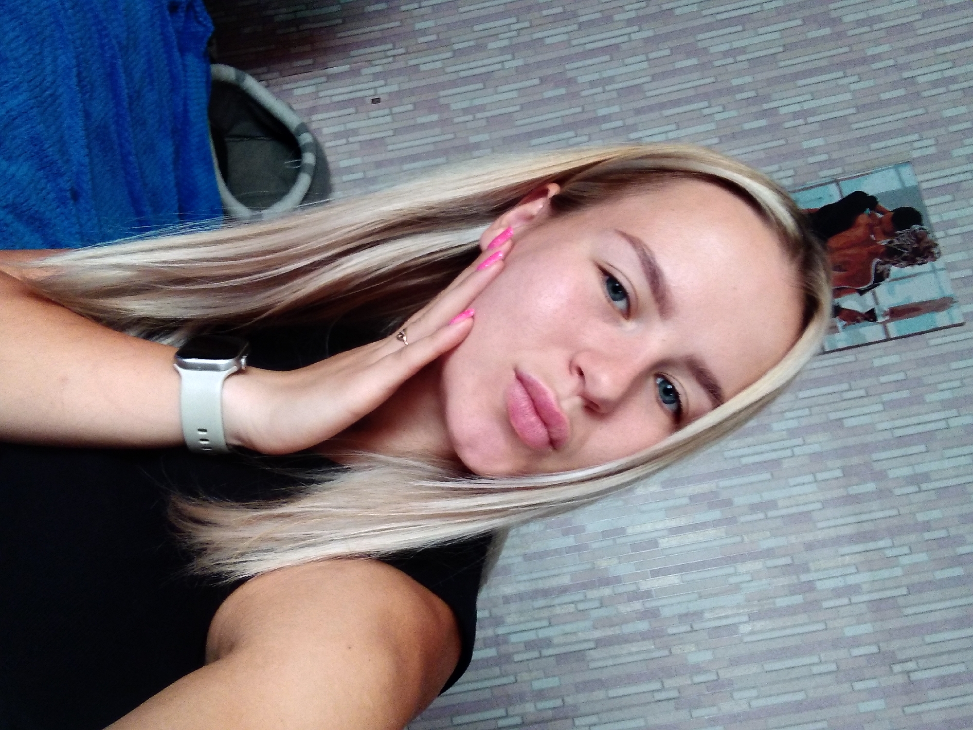 A young woman with blonde hair is taking a selfie in an indoor setting. She is wearing a black top and a smartwatch. Her hand is resting on her cheek, and her nails are painted bright pink. Behind her, the wall is covered with a patterned wallpaper. A framed picture of a couple is hanging on the wall above her head. A blue blanket and a gray bag are visible in the background. The overall lighting suggests the photo was taken during the daytime. The location is identified as Radisne, Ukraine.