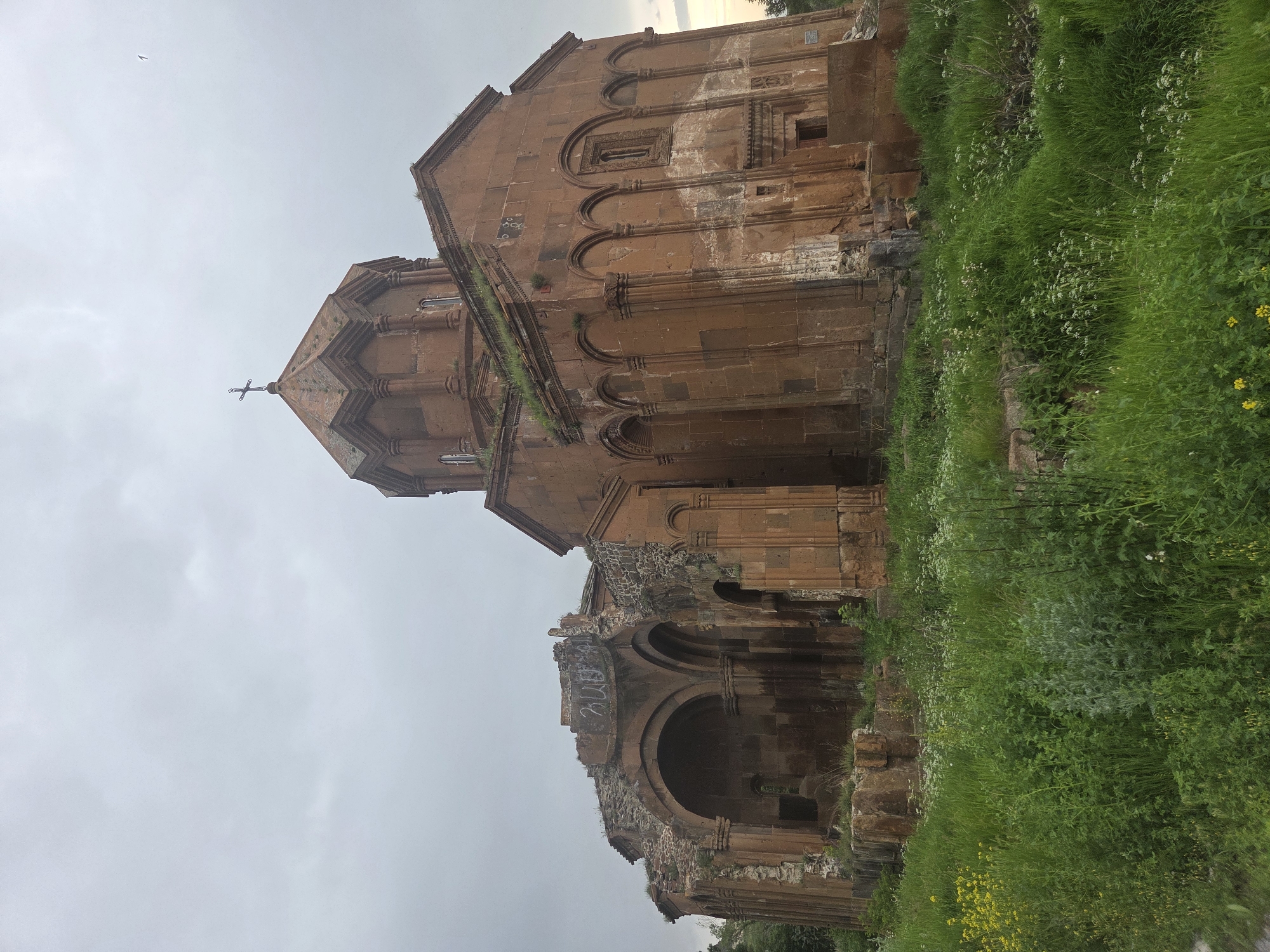 The image shows the semi-ruined medieval Armenian monastery of Talin, located in the village of Vahramaberd, Armenia. The structure is constructed from reddish-brown tuff stone and exhibits a central dome topped with a cross. The monastery appears overgrown with grass and vegetation, particularly around its base and on the roof. The left side of the building shows significant damage and crumbling walls, with arched openings visible. Graffiti is present on the upper part of the damaged section. The sky is overcast with a light gray hue. The foreground is filled with dense green grass and small wildflowers, including yellow and white varieties.