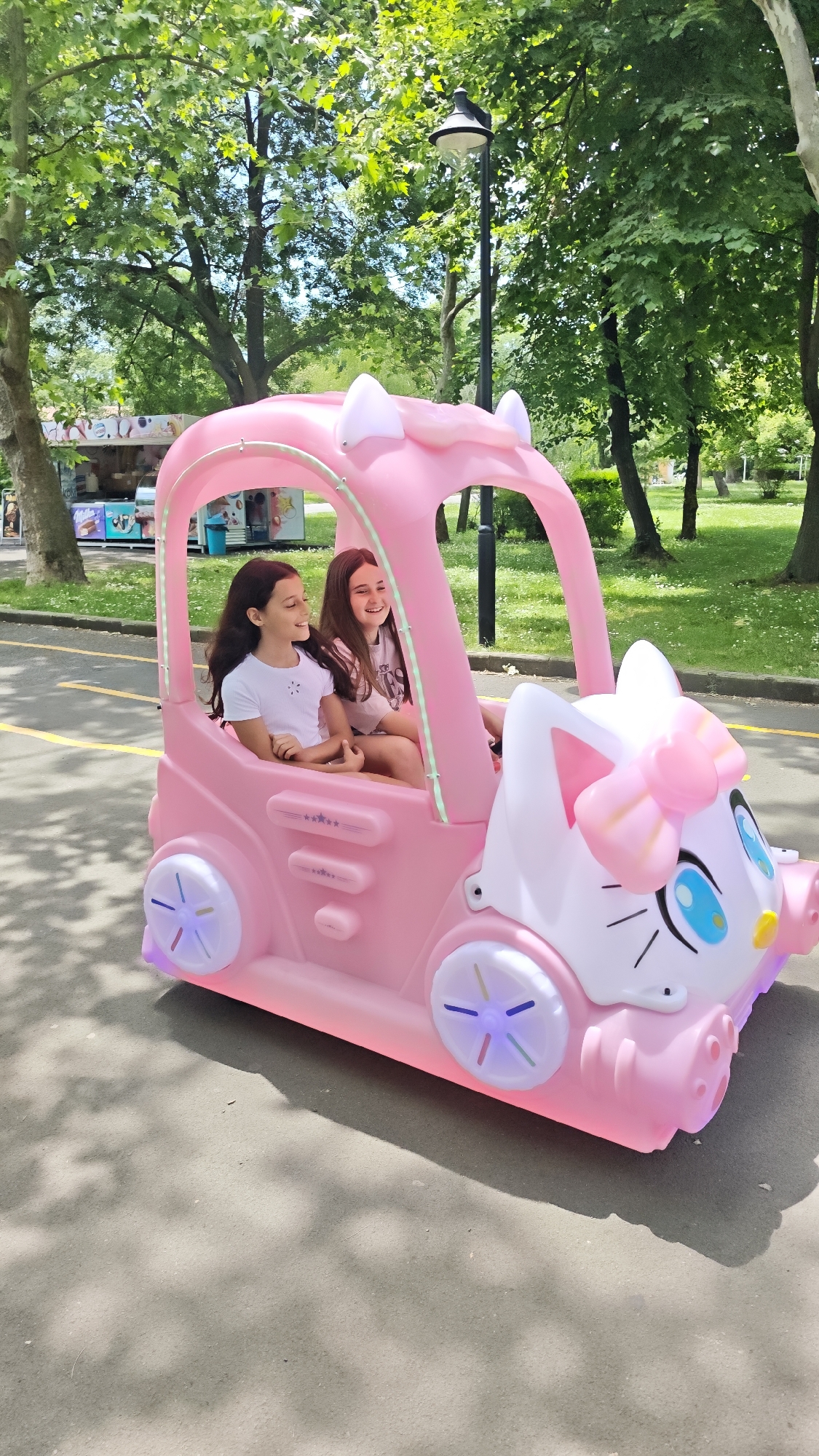 In a city park in Burgas, Bulgaria, two young girls, estimated to be between 8-12 years old, are riding in a pink, toy-sized, battery-powered car designed to resemble Hello Kitty. The car features illuminated wheels and a distinctive cat-like face. The girl on the left is wearing a white t-shirt and has dark hair. The girl on the right is wearing a light-colored t-shirt and also has dark hair. They are both smiling.
The setting includes a paved path, trees, and a nearby refreshment stand. A lamppost is also visible in the background. The overall atmosphere is sunny and cheerful.