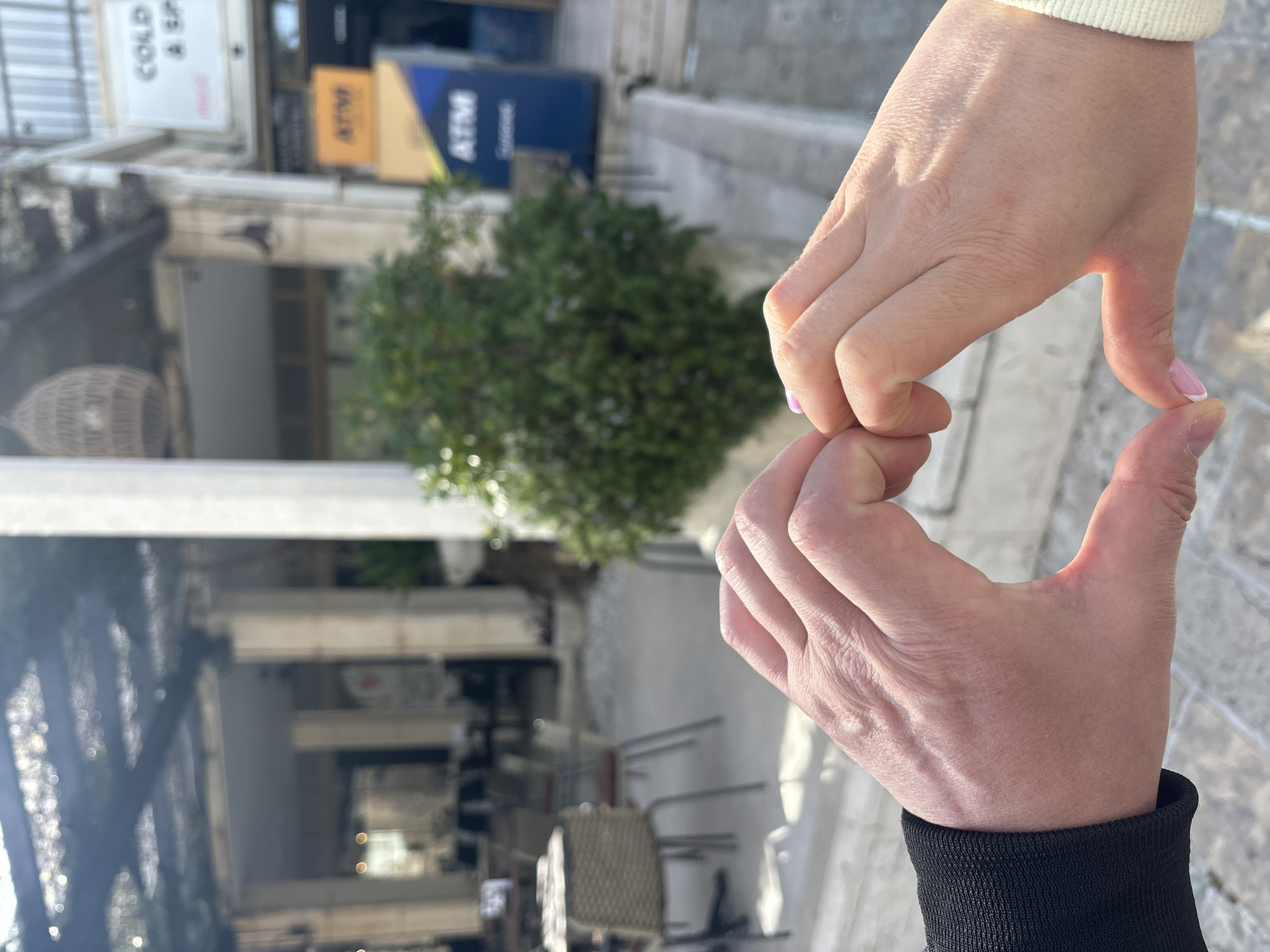 Two hands are forming the shape of a heart. The background includes a street scene in Budva, Montenegro. There is a building with a "COLD" sign and an ATM. A small green tree is also visible. The street has tables and chairs in front of the building.