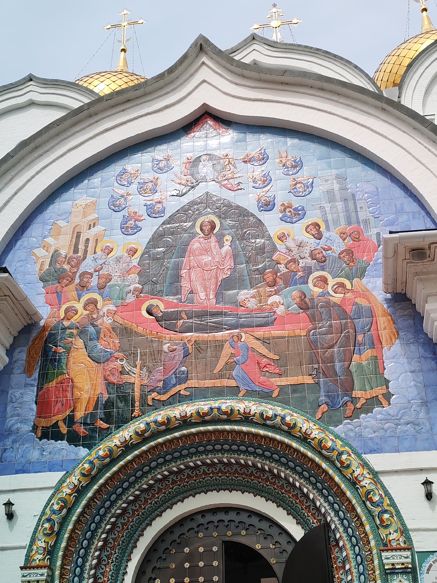 This image shows a colorful ceramic mosaic depicting a religious scene on the facade of a white building with a golden dome and a cross at the top. The mosaic, rendered in a style reminiscent of Orthodox icons, illustrates the burial of Christ.
In the center of the mosaic, Jesus is shown lying on a bier, dressed in a pink robe. A figure in a pink robe, presumably Jesus himself, stands to the right of the bier, blessing the body. Surrounding the bier are numerous figures, many with halos, representing mourners and holy individuals. Some are depicted with sorrowful expressions, leaning over the body or looking on with solemnity. Angels are also present, some in the sky above and others near the figures on the ground. In the background, there are architectural elements resembling buildings or perhaps a depiction of the celestial realm. The mosaic is set within an arched architectural feature of the building, which is further decorated with intricate, colorful patterns. The overall impression is one of solemnity and reverence, typical of religious iconography. The building's architecture, with its golden dome and crosses, suggests it is a church or a similar religious structure, likely located in Russia given the context provided. The sky appears overcast, suggesting it could be daytime with diffused light.
