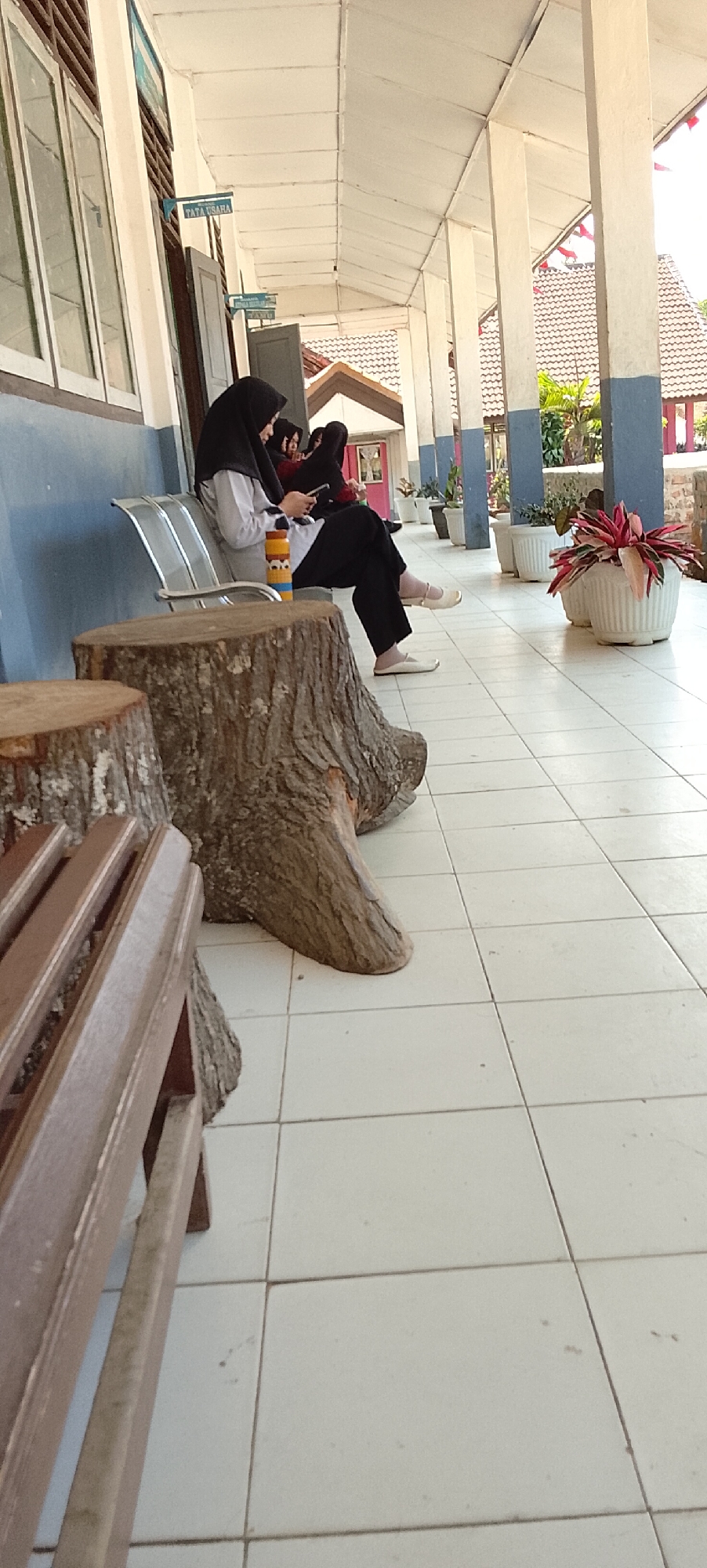 The image captures a scene on a tiled veranda, possibly at an educational institution in Jepara, Indonesia, indicated by a sign that reads "TATA USAHA." Three individuals, two women and one girl, are seated on a metallic bench. The woman in the foreground, wearing a black hijab and light-colored clothing, is looking down at her phone. Beside her, another woman, also in a black hijab, is partially visible. A young girl, seated at the far end of the bench, is partially obscured and appears to be looking in the same direction as the first woman.
In front of the bench are two large, rough-hewn tree stumps, serving as makeshift seats or decorative elements. The veranda is supported by white pillars with blue accents at the base, and the ceiling is white and paneled. The tiled floor is light-colored and grid-patterned.
Further down the veranda, several white planters contain lush green and red foliage. In the background, the roof of a building with a reddish-brown tile pattern is visible, along with the suggestion of trees and other vegetation. The overall atmosphere suggests a relaxed moment during the daytime, possibly within a school or office complex.