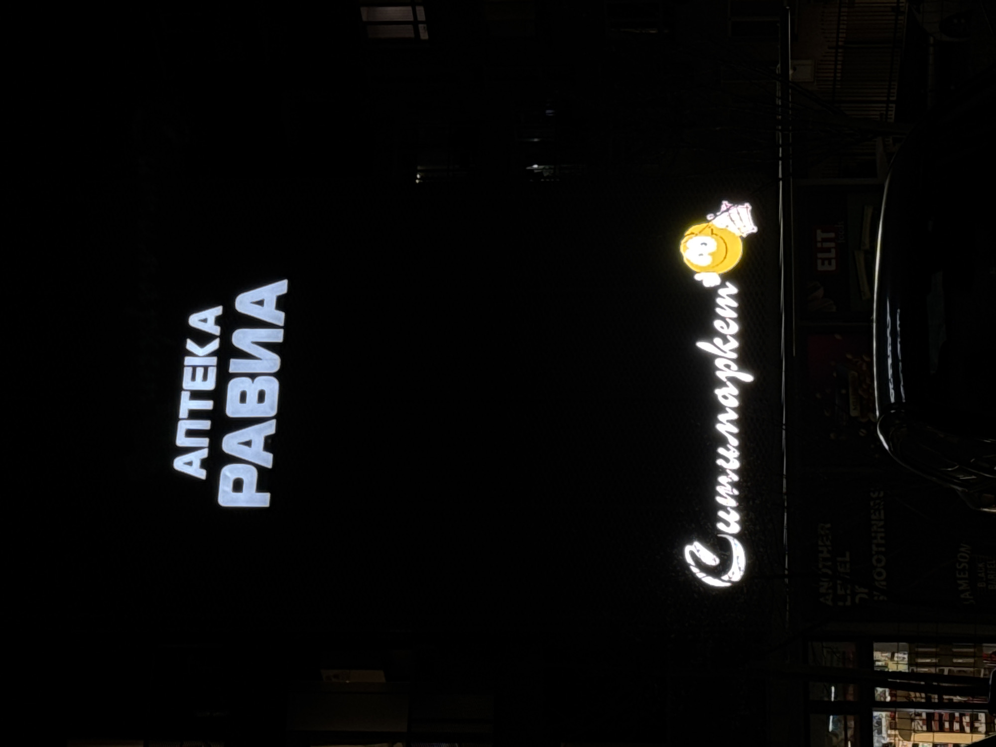 At night, the image depicts illuminated signs in Varna, Bulgaria. On the left is the word "АПТЕКА" above "РАВИА" in white block letters. To the right, another illuminated sign reads "Cumu.napkem" in cursive, accompanied by a yellow cartoon face. Adjacent to this is a sign for "ELIT". Below the cursive sign, partially visible signage includes the words "ANOTHER LEVEL OF SMOOTHNESS" and "JAMESON BLACK BARREL". The area appears to be a business district, with a building facade visible behind the signs.