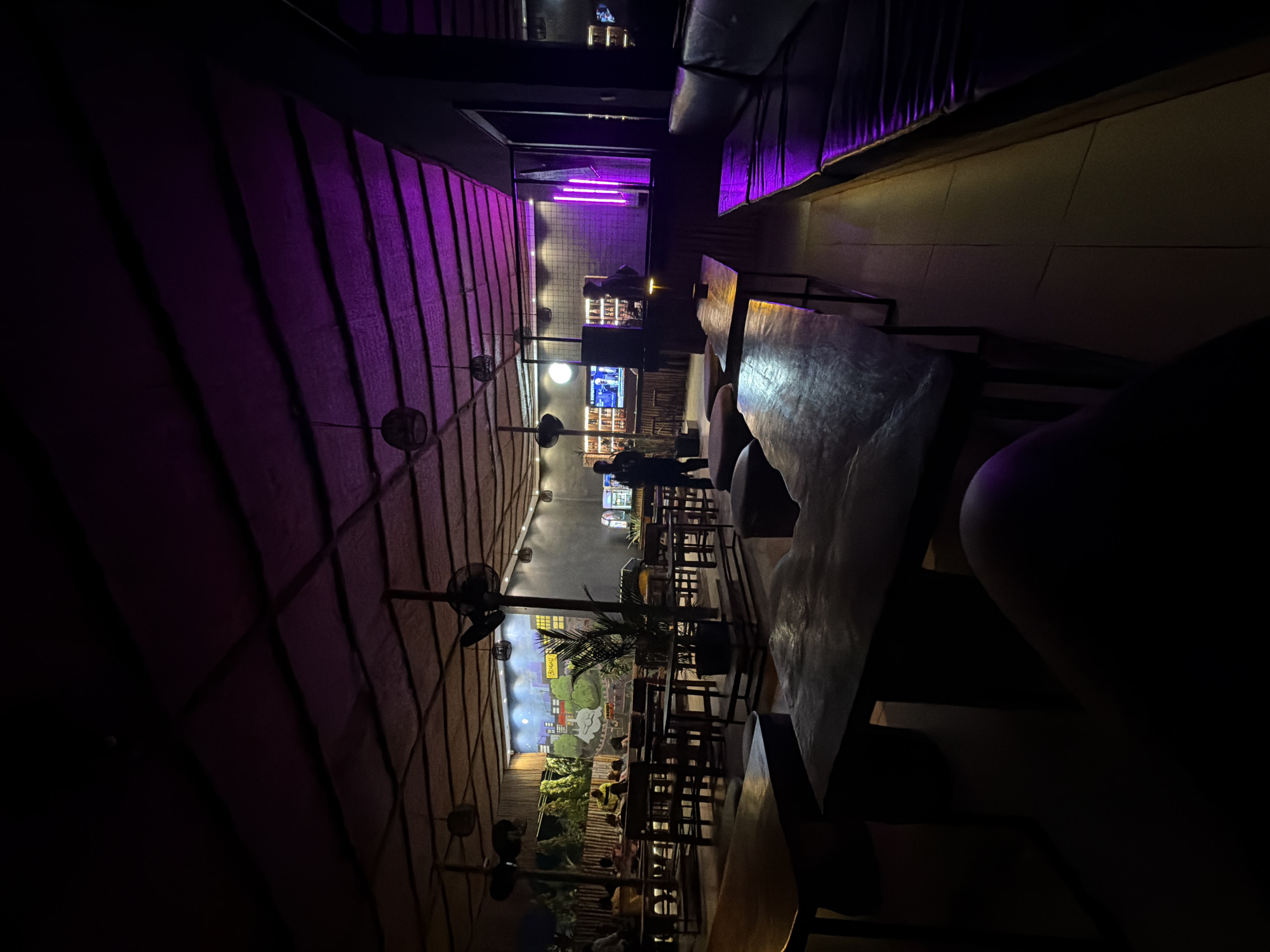 The image depicts the interior of a dimly lit establishment, likely a restaurant or bar, in Gwarinpa, Nigeria. The ceiling is constructed from woven materials, with exposed rafters crisscrossing overhead. A purple hue from ambient lighting casts a glow on portions of the ceiling and illuminates a black leather sofa on the right side of the frame. In the background, a mural or painted wall displays a stylized cityscape with trees and buildings. Several wooden tables and benches are arranged throughout the space, some occupied by patrons who are not clearly visible due to low light conditions. A bar area is visible in the middle ground, with shelves stocked with bottles. Two individuals, silhouettes against the background lighting, stand near the bar. The foreground is dominated by a dark wooden table and bench, with a person's leg visible in the lower left corner, also silhouetted. The overall ambiance suggests a nighttime setting with artificial lighting creating a mood.