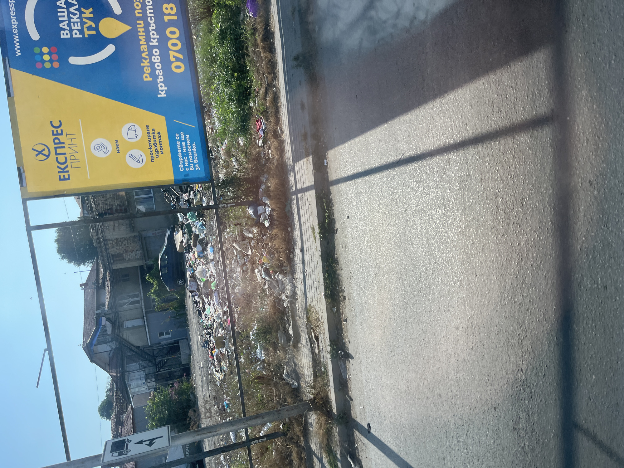 The image depicts a neglected urban area with a significant amount of trash strewn across the ground. In the background, a multi-story building with peeling paint and a metal fire escape is visible. Adjacent to it is a billboard advertising a printing and advertising service. The billboard is predominantly yellow and blue and features text in Bulgarian. The lower portion of the image shows a street or paved area with tire tracks and shadows cast by an unseen object, likely a vehicle. The scene appears to be located in Varna, Bulgaria, as indicated by the billboard's language and general aesthetic. The overall impression is one of urban decay and disregard for public space.