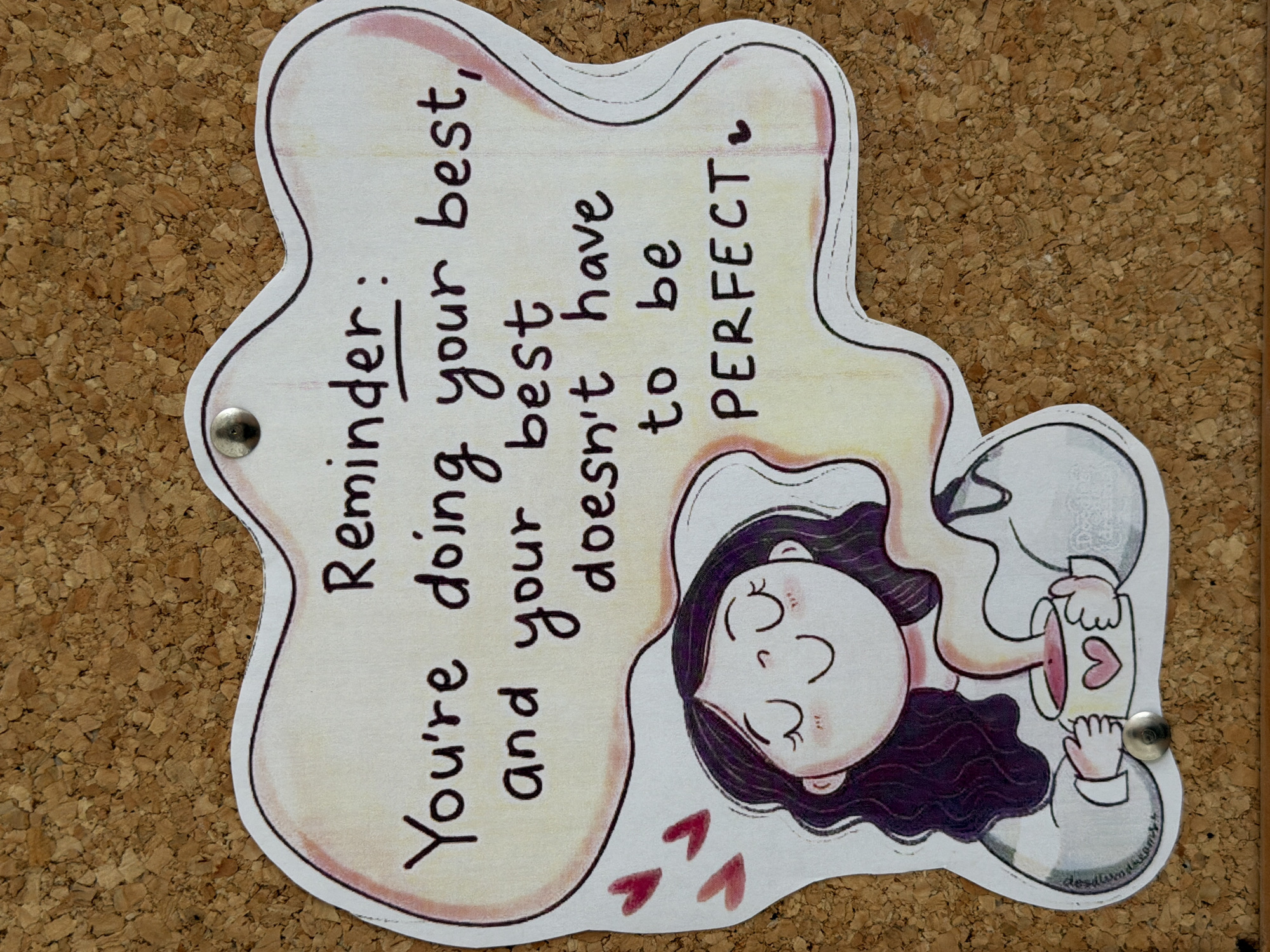 The image shows a reminder note pinned to a corkboard. The note is shaped irregularly, with a white background and a thin black outline. It reads "Reminder: You're doing your best, and your best doesn't have to be PERFECT". Below the text, there is a cartoon illustration of a person lying down, eyes closed, holding a mug with a heart on it. The person has dark hair and is wearing a light-colored shirt. There are small heart shapes floating around the person. The note is attached to the corkboard with two silver pushpins.